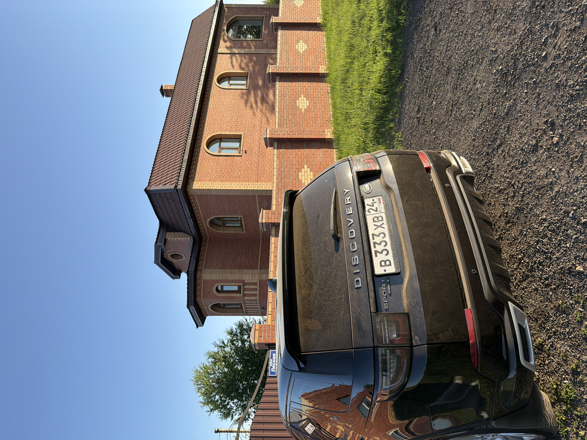 A black Land Rover Discovery Sport SUV is prominently featured, parked on a gravel path next to a large red brick house under a clear, bright blue sky. The scene is illuminated by strong sunlight, suggesting it is midday or early afternoon.
The rear of the SUV, which appears somewhat dusty, faces the viewer. Visible text on the vehicle includes "DISCOVERY SPORT" on the tailgate and the Russian license plate "B 333 XB 24 RUS." The license plate frame carries additional text in Cyrillic: "ВЕСТИАНСТЕР СЕРВИС MINOR JAGUAR LAND," likely identifying a dealership.
The brick house, standing to the left and behind the car, has a dark tiled roof and features several arched windows and decorative brick patterns. Green grass lines the edge of the gravel path, separating it from the house. A leafy green tree is partially visible in the mid-ground. A blue street sign, oriented vertically, displays the Cyrillic word "Лесная" (Lesnaya), which translates to "Forest" or "Wooded." This combination of a Russian license plate and a street sign with Cyrillic text, in conjunction with the provided context, places the scene in Krasnoyarsk, Russia. No people or specific activities are visible in the static composition.
