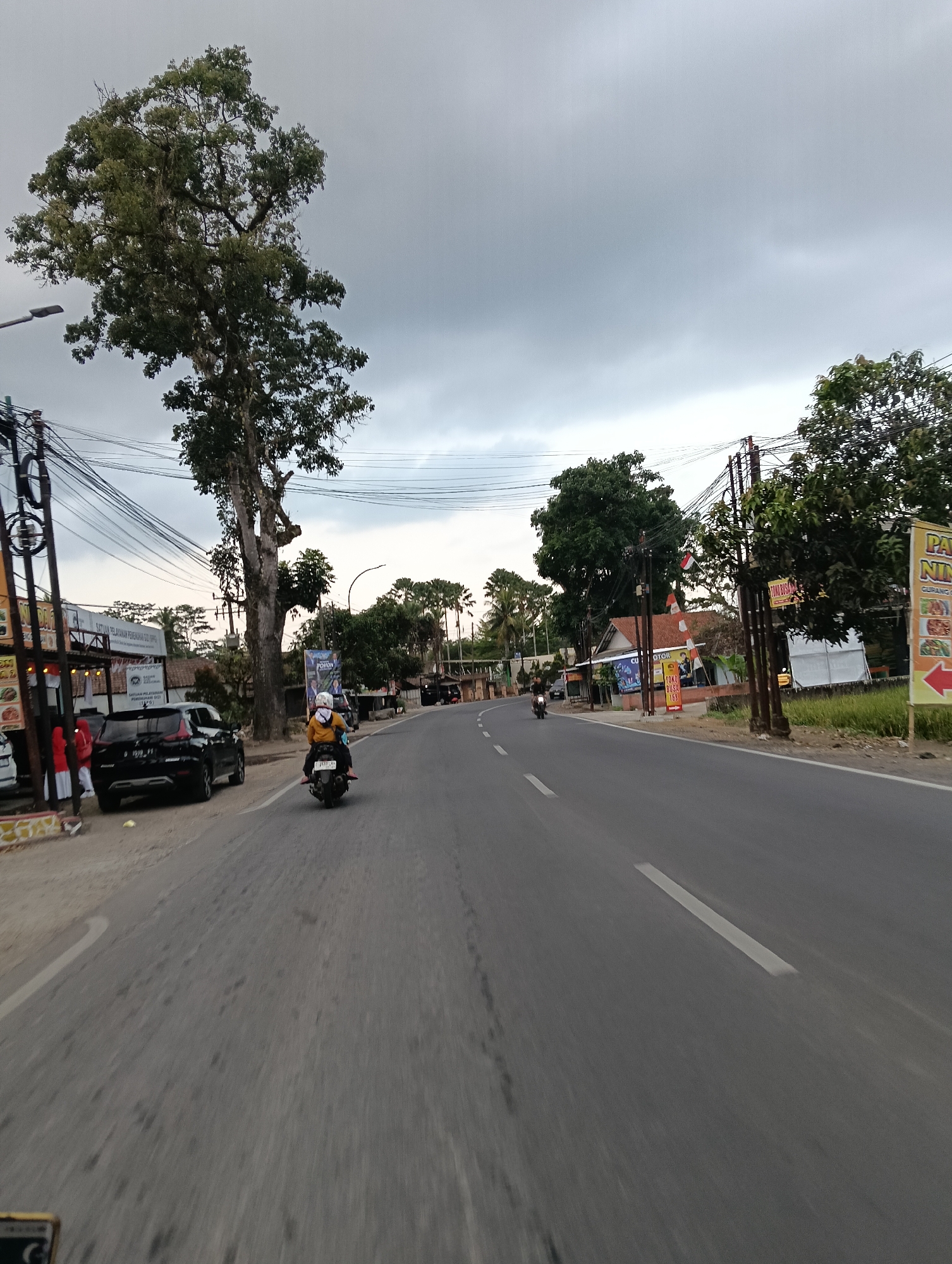 The image captures a street scene in Cintaraja, Indonesia, with a focus on a paved road under a cloudy sky. A large, mature tree with dense foliage dominates the left side of the frame, its branches reaching towards the sky. Power lines crisscross overhead, supported by poles along the roadside. On the left, a black SUV is parked next to a building with an awning. People are visible near the building, including one in red attire. Further down the road, a person on a motorcycle is riding away from the camera, wearing a yellow jacket and a white helmet. The road has white lane markings. On the right side of the road, there are more trees and buildings, with signs and what appears to be a rice paddy field. A yellow banner with red text and an arrow is prominently displayed on the right. The overall impression is of a semi-urban or rural environment, possibly in the late afternoon given the lighting.