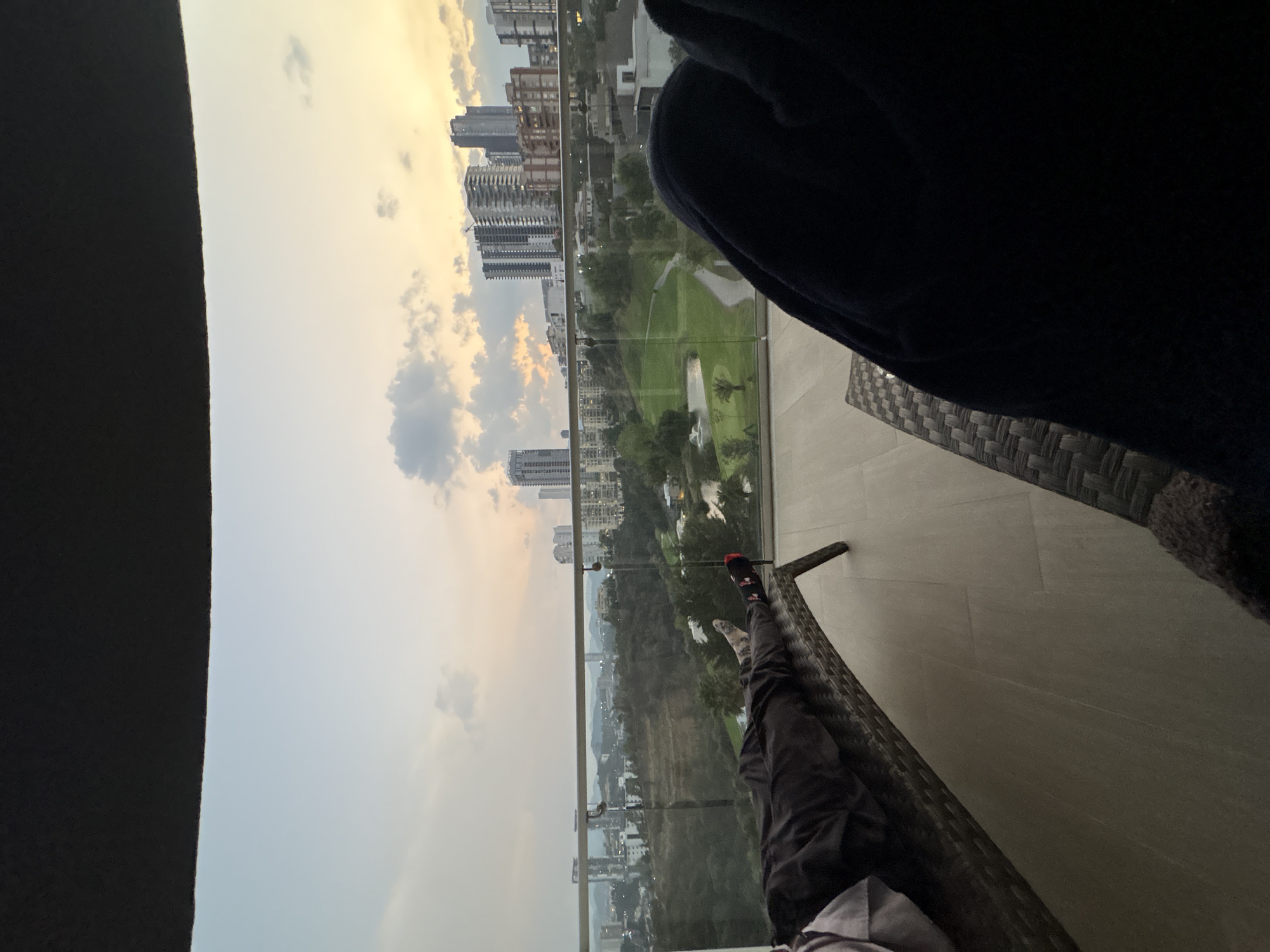 The image depicts a person relaxing on a balcony, likely in a high-rise building, with a view overlooking a cityscape and a golf course. The person is wearing dark pants and socks, one of which is red and black. They are seated on a wicker chair. A dark-colored blanket or garment is draped nearby.
Beyond the balcony railing, the view includes numerous buildings in the city of Huixquilucan de Degollado, Mexico. A golf course with green fairways and water features is visible below. The sky is filled with clouds, suggesting either sunrise or sunset.