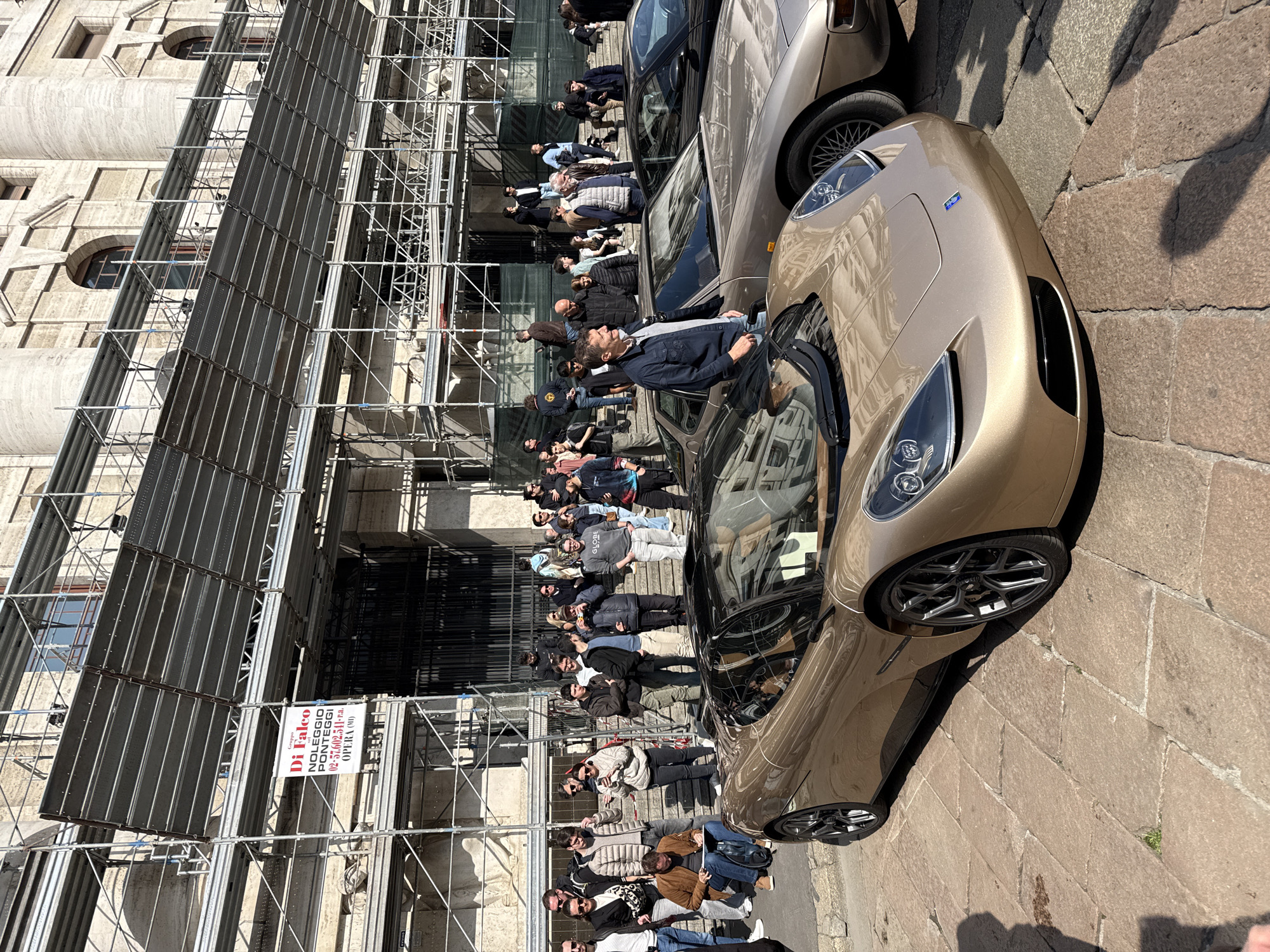 The image depicts an outdoor scene in Piazza Affari, Milan, Italy, in front of the Palazzo della Borsa, which is undergoing renovation. A large, ornate neoclassical stone building forms the background, almost entirely obscured by extensive silver-colored metal scaffolding. A white banner with red text, identifying "Gruppo Di Falco NOLEGGIO PONTEGGI" and contact information, hangs from the scaffolding.
In the foreground and midground, two high-performance sports cars are parked on a light-colored cobblestone street. The car closest to the viewer is a modern, champagne-bronze colored vehicle with sleek lines, a black roof, and dark multi-spoke wheels; a small blue emblem is visible on its front bumper. Immediately behind it, a vintage champagne-bronze sports car, likely a Porsche model, is visible with distinctive lattice-style wheels. A third, dark-colored car is partially visible behind the vintage car.
A crowd of approximately 30-40 individuals is gathered around the vehicles and on the wide stone steps leading up to the building's entrance. Most individuals are casually dressed. A man in a dark blue jacket and light blue jeans stands beside the vintage car, looking to his right. Another man, wearing a brown jacket, is seated on a low structure to the left of the modern car, engaged in sketching on a pad. Numerous other individuals are standing, observing the cars, conversing, or using smartphones, some appearing to photograph the scene. The scene is illuminated by bright sunlight, casting distinct shadows on the cobblestone ground.