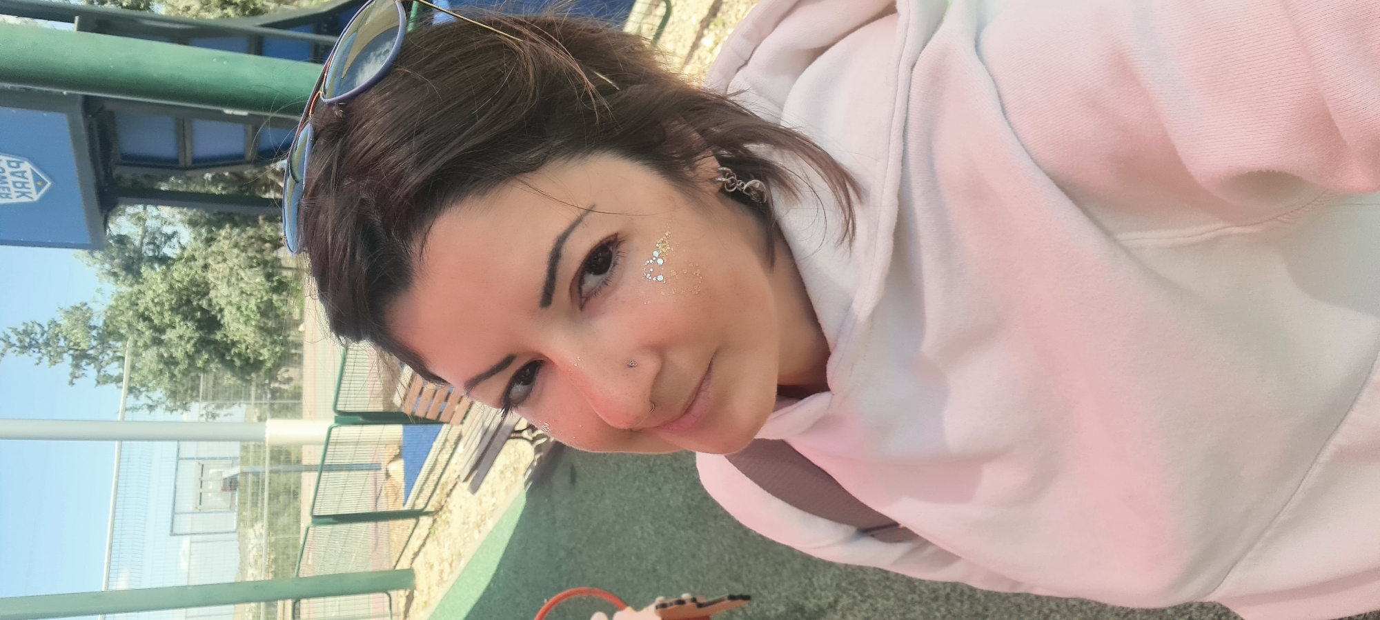 A woman with glitter on her face takes a selfie. She is wearing a white hoodie and has sunglasses perched on her head. She has a nose ring and earring. A sign for a park is visible behind her, along with trees, a fence, and a basketball court. The location is in the city of Lachish Regional Council, Israel.