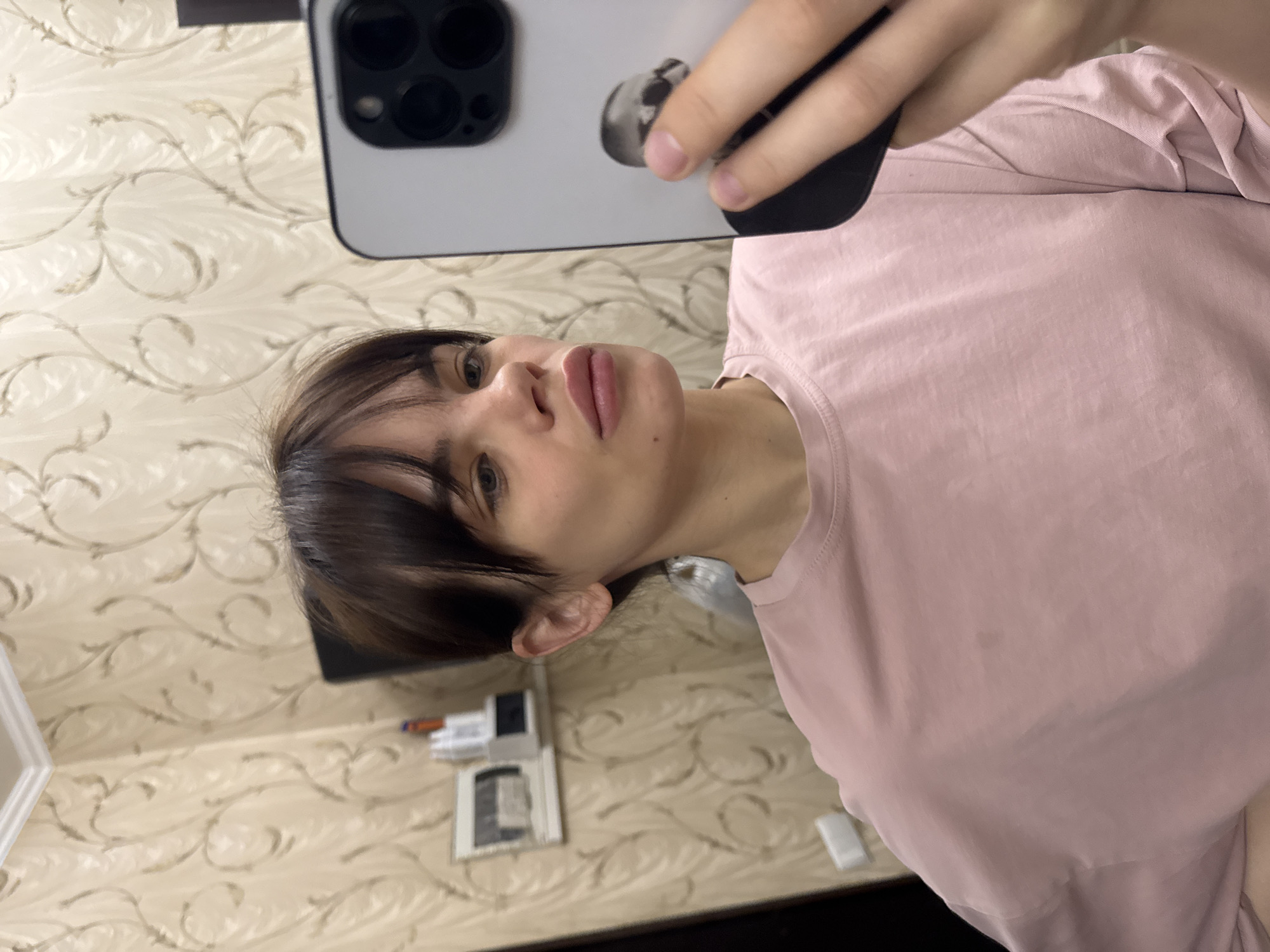 A woman with brown hair and bangs, wearing a pink t-shirt, is taking a selfie with a white smartphone. The phone's camera lens is visible. The background is a wall with a patterned wallpaper. A small white frame and what appears to be a child's toy are visible on the wall. The location appears to be an indoor space in Rostov-on-Don, Russia.