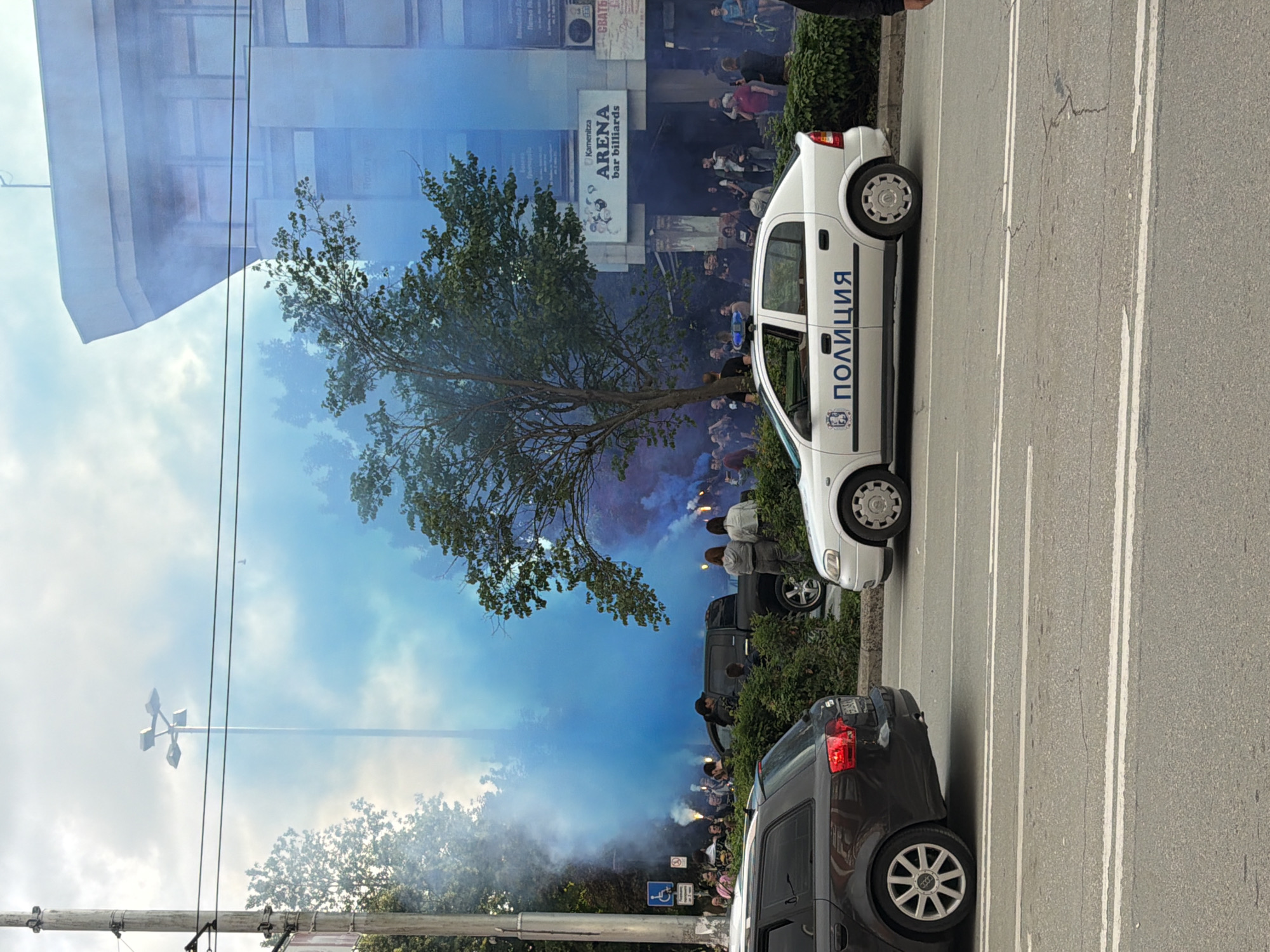 A street scene in Pleven, Bulgaria shows a crowd of people holding flares and emitting blue smoke in front of a building with a sign "ARENA bar billiards." A white police car, labeled "ПОЛИЦИЯ" (Police) in Cyrillic script, is parked on the street. Two other cars, one dark colored, are also parked nearby. The scene seems to be a gathering or demonstration. The street is paved and marked with lane dividers. The sky is visible through the smoke and the reflection of the building. A street lamp pole is also visible.