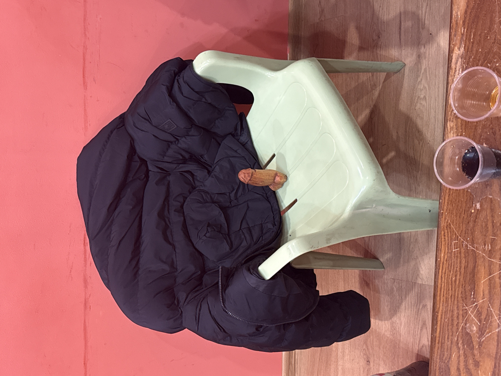 A black puffer jacket is draped over a light green plastic chair next to a wooden table. Two clear plastic cups, one with a dark substance and the other with a yellowish liquid, are on the table. The setting is indoors, with a reddish-pink wall visible behind the chair and jacket. The floor appears to be laminate wood. The location is indicated as Sofia, Bulgaria.
