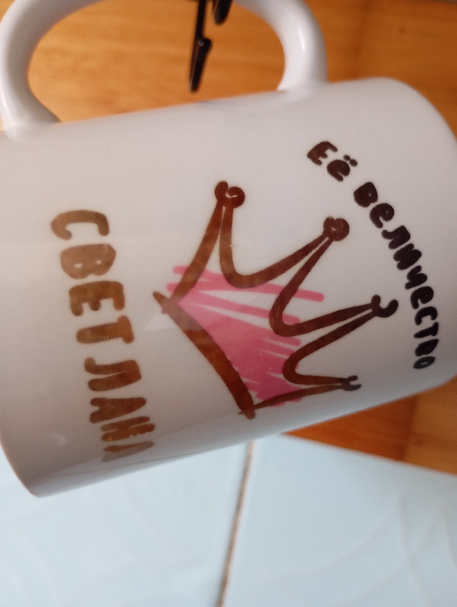 A close-up, angled shot captures a white ceramic mug adorned with brown lettering and a drawing of a crown. The lettering on the left side of the mug reads "СВЕТЛАНА" vertically, and on the right side, it reads "её величество" in a curve. The crown drawing features a pink fill within its points. The mug is held at an angle, with its handle visible at the top. The background consists of a wooden surface and a light blue surface, suggesting it might be placed on a table with a wooden tabletop or shelf. The image appears to be taken in Gorki-2, Russia, judging by the provided context. There are no people or explicit actions depicted in the image.