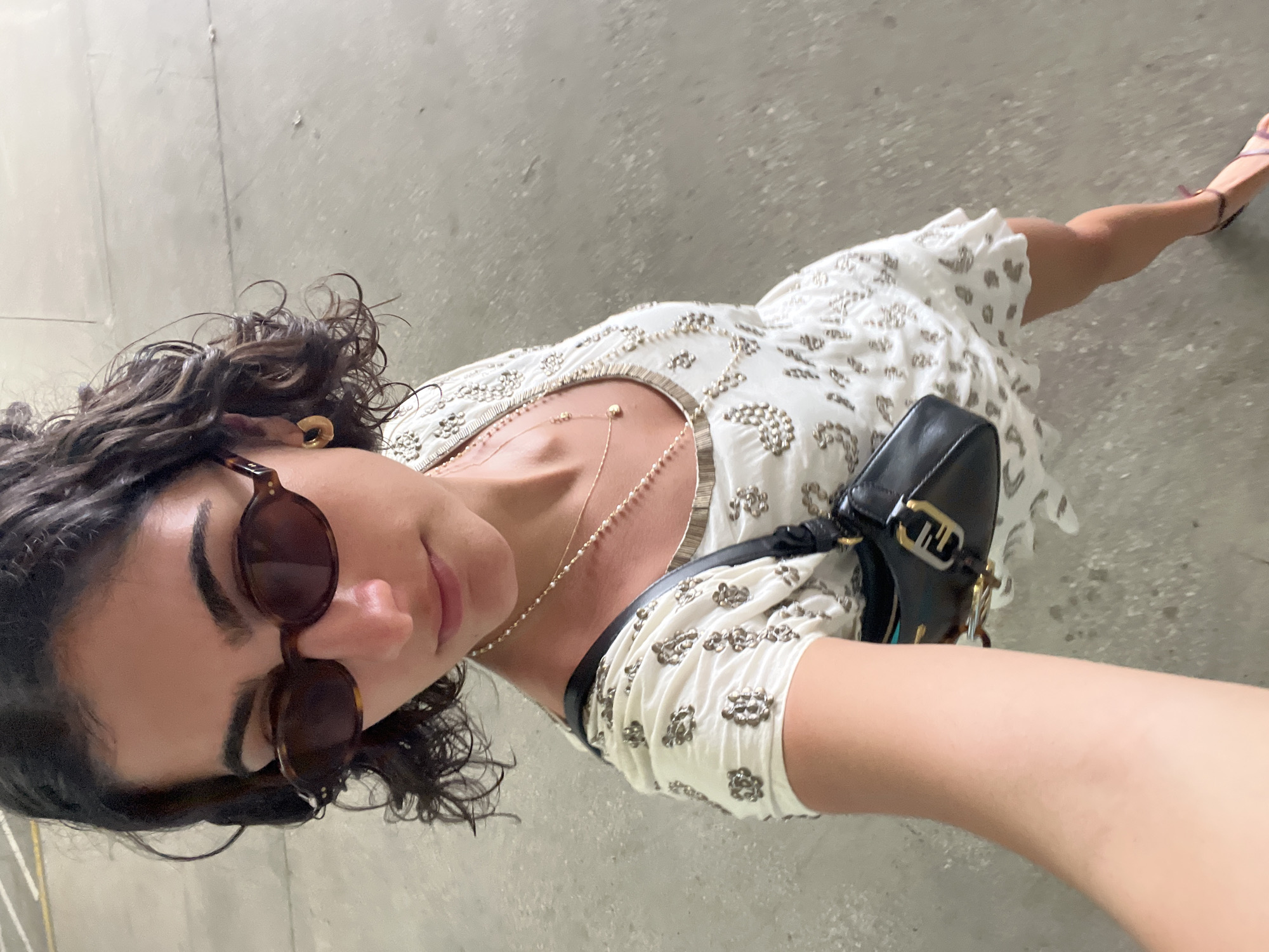 A young woman is standing on a gray concrete surface. She is wearing sunglasses and a white dress with silver embellishments. She is carrying a black leather purse. She has curly dark hair and is wearing gold earrings. The location is likely outdoors, but the specific details of the city of Miami, United States of America, cannot be determined from the provided image.