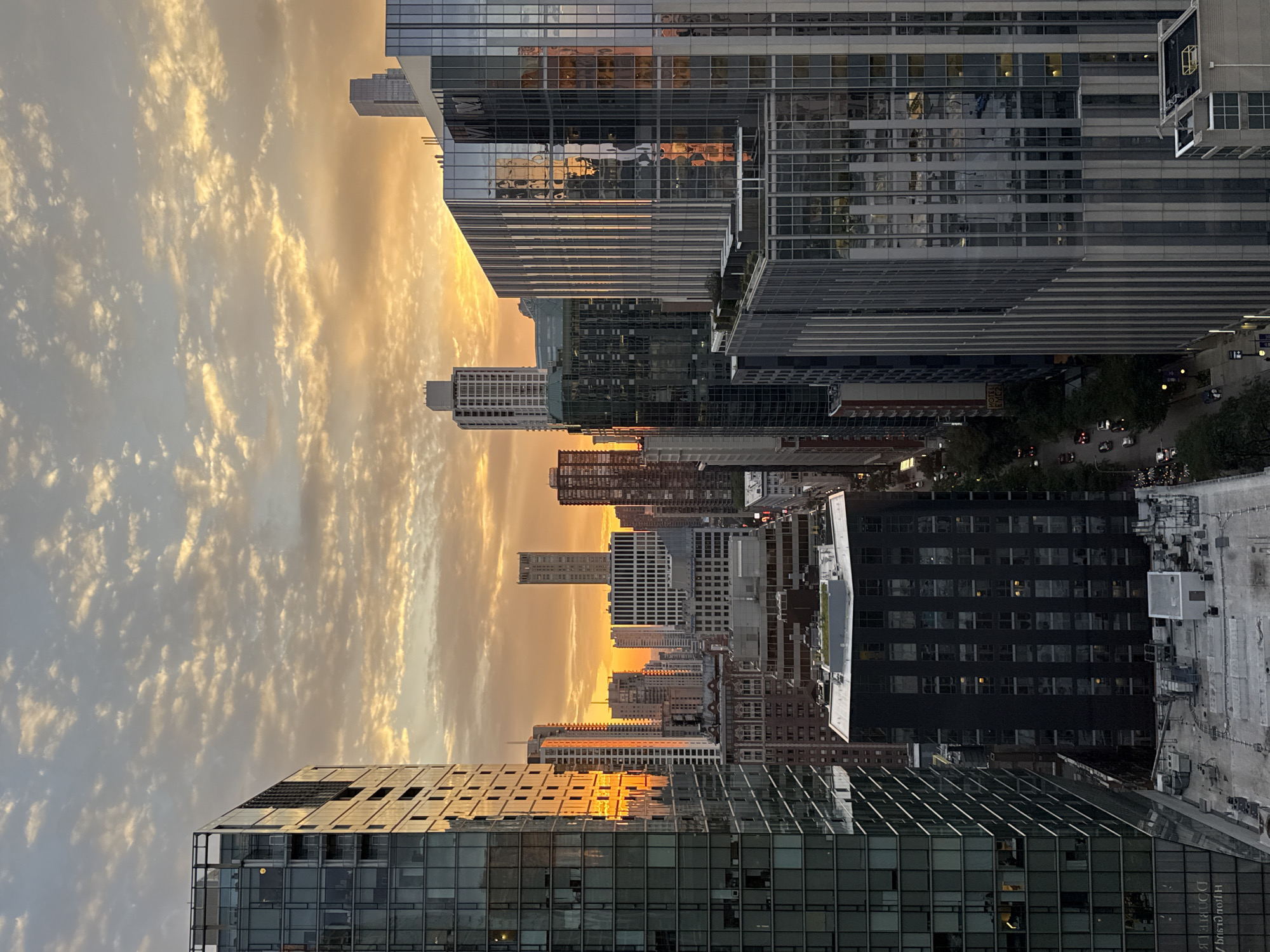 This is a photograph of a city skyline at sunset. The sky is filled with clouds that are illuminated by the setting sun, casting an orange and yellow glow. Numerous skyscrapers of varying heights and architectural styles dominate the foreground and midground. The buildings are made of glass and concrete, with many featuring reflective surfaces that mirror the colors of the sky. Some buildings appear to be residential, with visible windows and balconies, while others are more commercial in appearance. The view is from an elevated perspective, suggesting the photograph was taken from a high floor of a building. The street level is visible in some areas, showing cars and trees, indicating an urban environment. The overall impression is one of a vibrant, modern city bathed in the warm light of sunset. The architecture and density of the buildings suggest a location like Chicago, a major metropolitan area known for its impressive skyline.