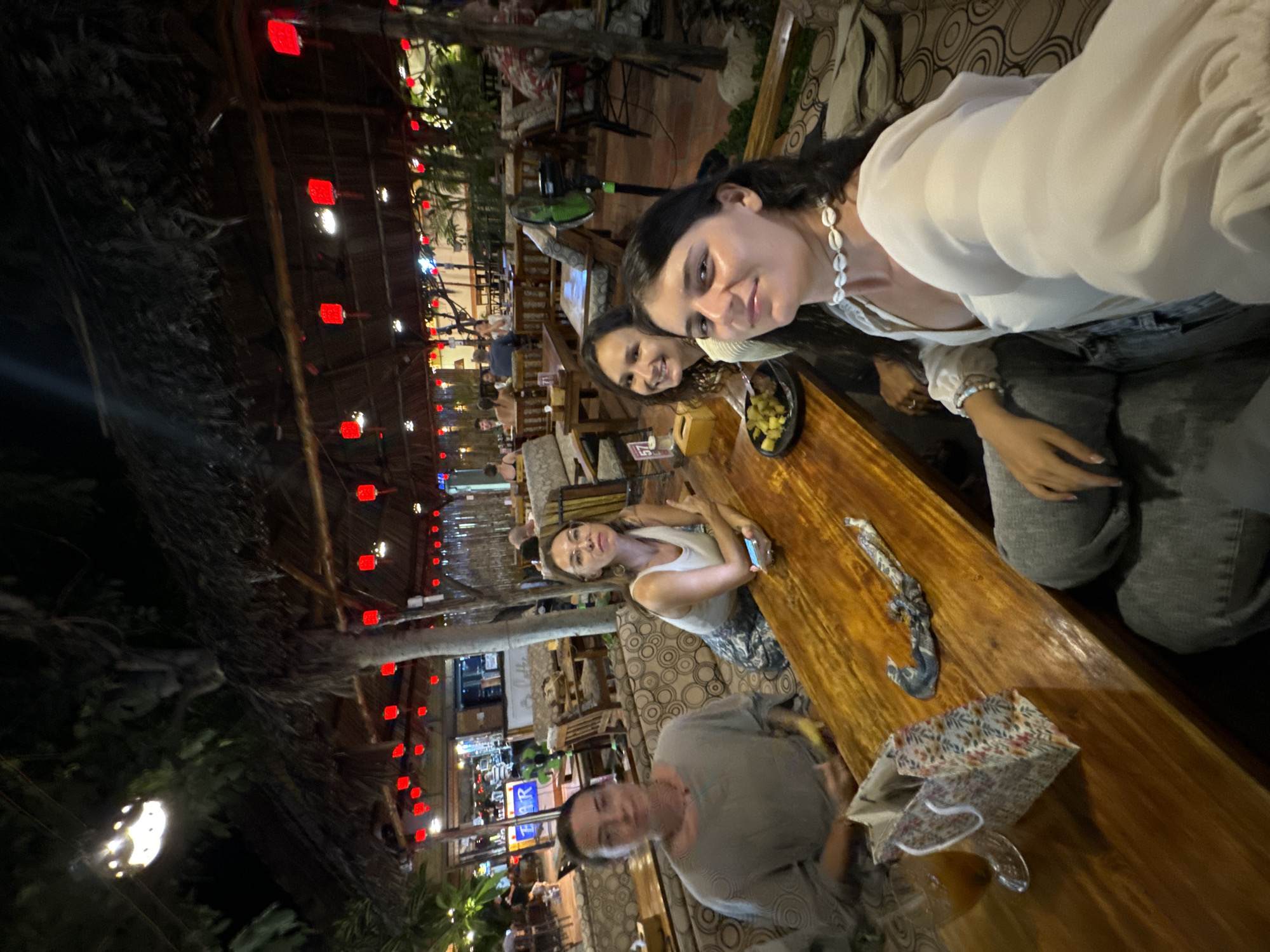 Four women are seated at a wooden table in an outdoor restaurant setting. The restaurant has a thatched roof, and is decorated with red lanterns. The women are dressed casually. The woman on the far left is wearing a grey top, the woman next to her is wearing a white tank top and a patterned skirt. The woman in the middle is wearing a black top, and the woman on the right is wearing a white blouse and a shell necklace. There is food on the table, as well as a gift bag and a glass of drink. The setting appears to be nighttime, with artificial lighting illuminating the scene. The restaurant is furnished with wooden tables and chairs, and cushioned seating. The background shows other patrons and tables, suggesting a lively atmosphere. The location is identified as Phu Thuy Ward, Vietnam.