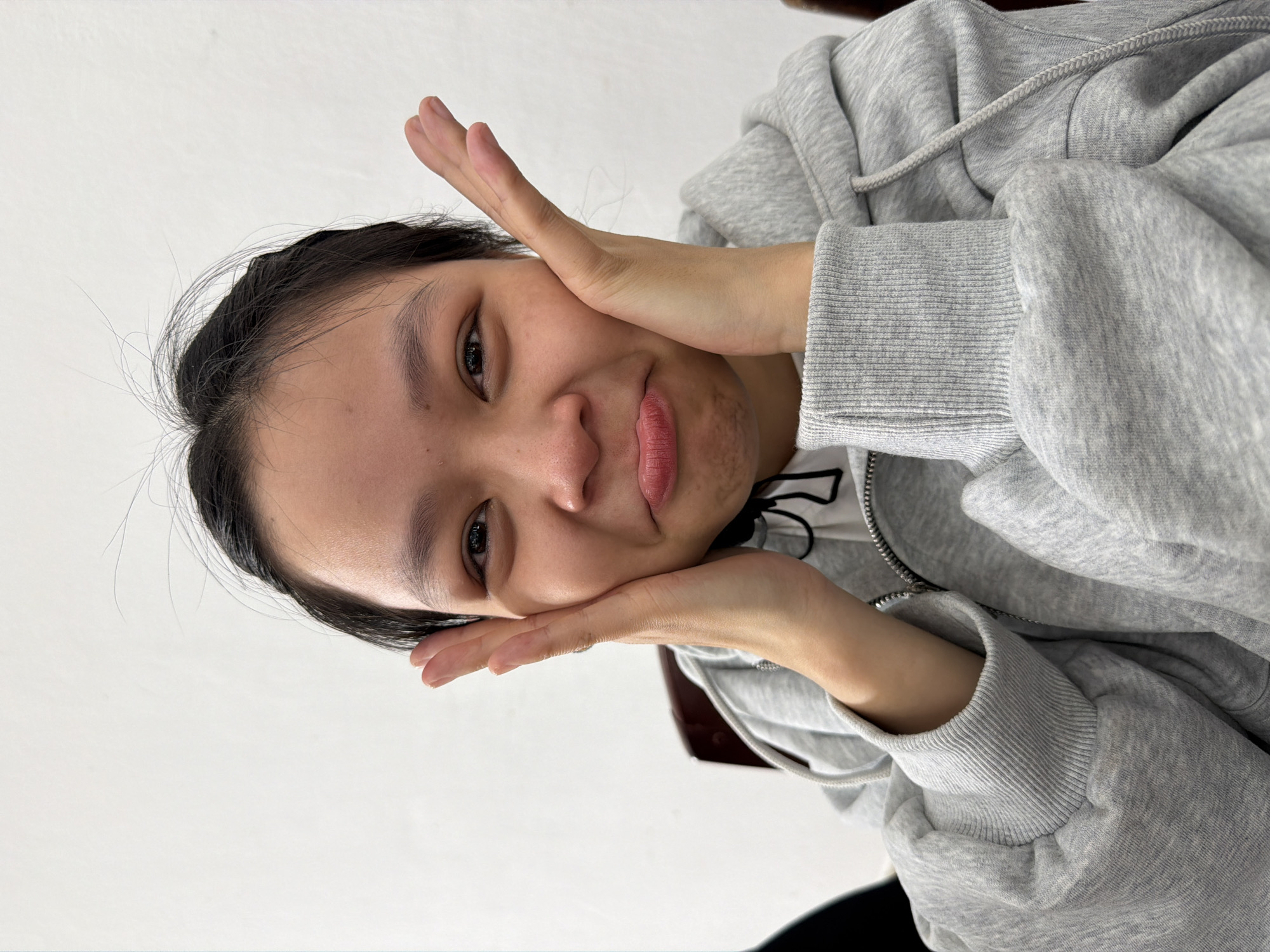 A young woman with dark hair pulled back from her face is seated, wearing a gray hooded sweatshirt. She is holding her hands on either side of her face, palms inward, as if making a "cute" pose. The background is a plain white wall. The location, Đà Nẵng, Vietnam, cannot be confirmed from the image.