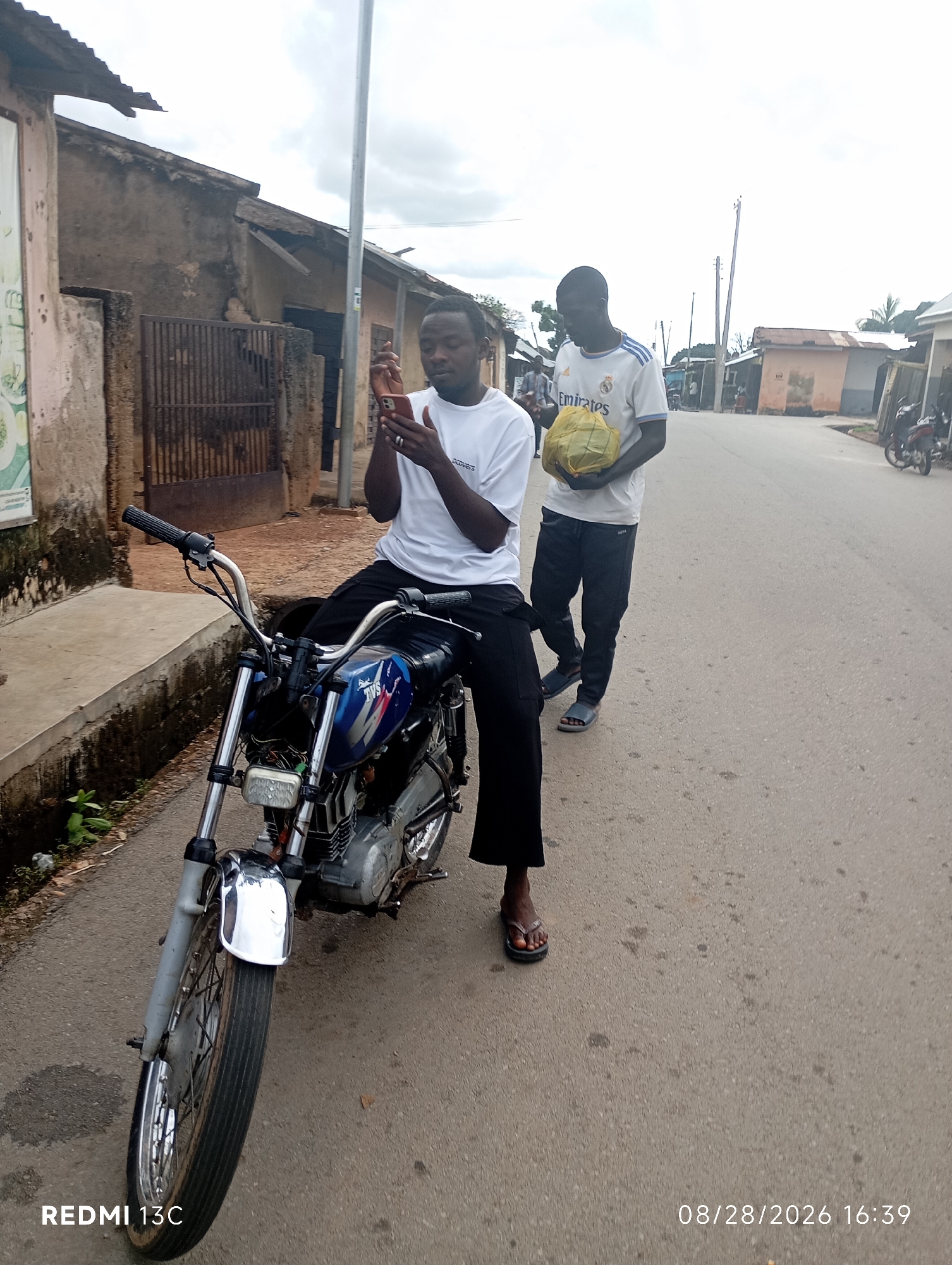 Two young men are present on an unpaved road in Shendam, Nigeria. The man on the left, wearing a white t-shirt and black trousers, is seated on a blue and silver motorcycle. He is holding a smartphone in his right hand and appears to be looking at it. The man on the right, wearing a white football jersey with "Emirates" printed on it and dark trousers, is standing next to the motorcycle. He is holding a yellow object that resembles a ball or a fruit in both hands. The background consists of modest buildings with corrugated iron roofs and a cloudy sky. The scene is captured with a timestamp of "08/28/2026 16:39" and the device "REDMI 13C" is indicated.