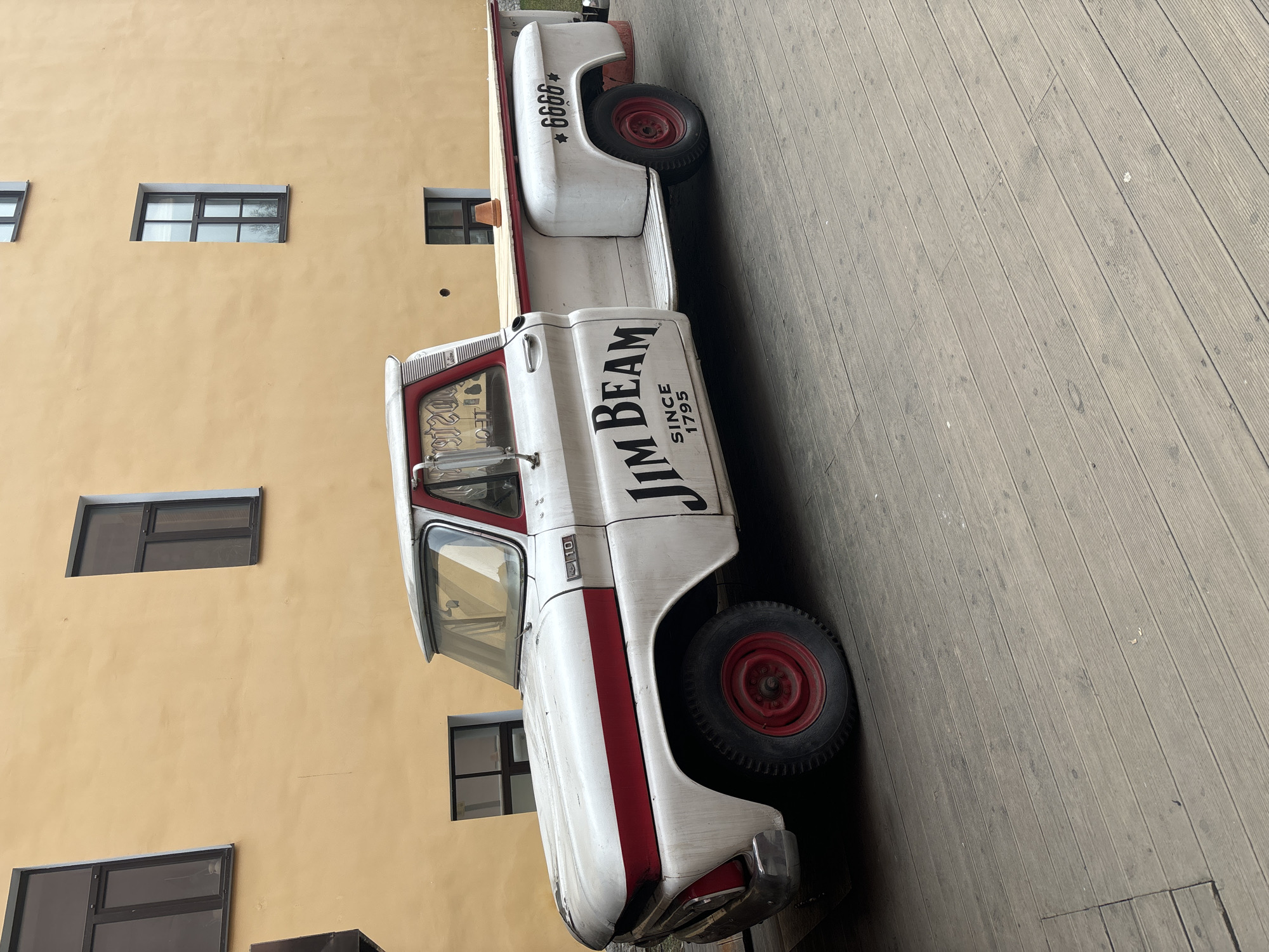 The image depicts a vintage white and red pickup truck parked on a wooden boardwalk next to a building with yellowish-beige walls and windows. The truck has "JIM BEAM SINCE 1795" written on its side. The number "6666" is visible on the rear of the truck. The location appears to be Saint Petersburg, Russia.