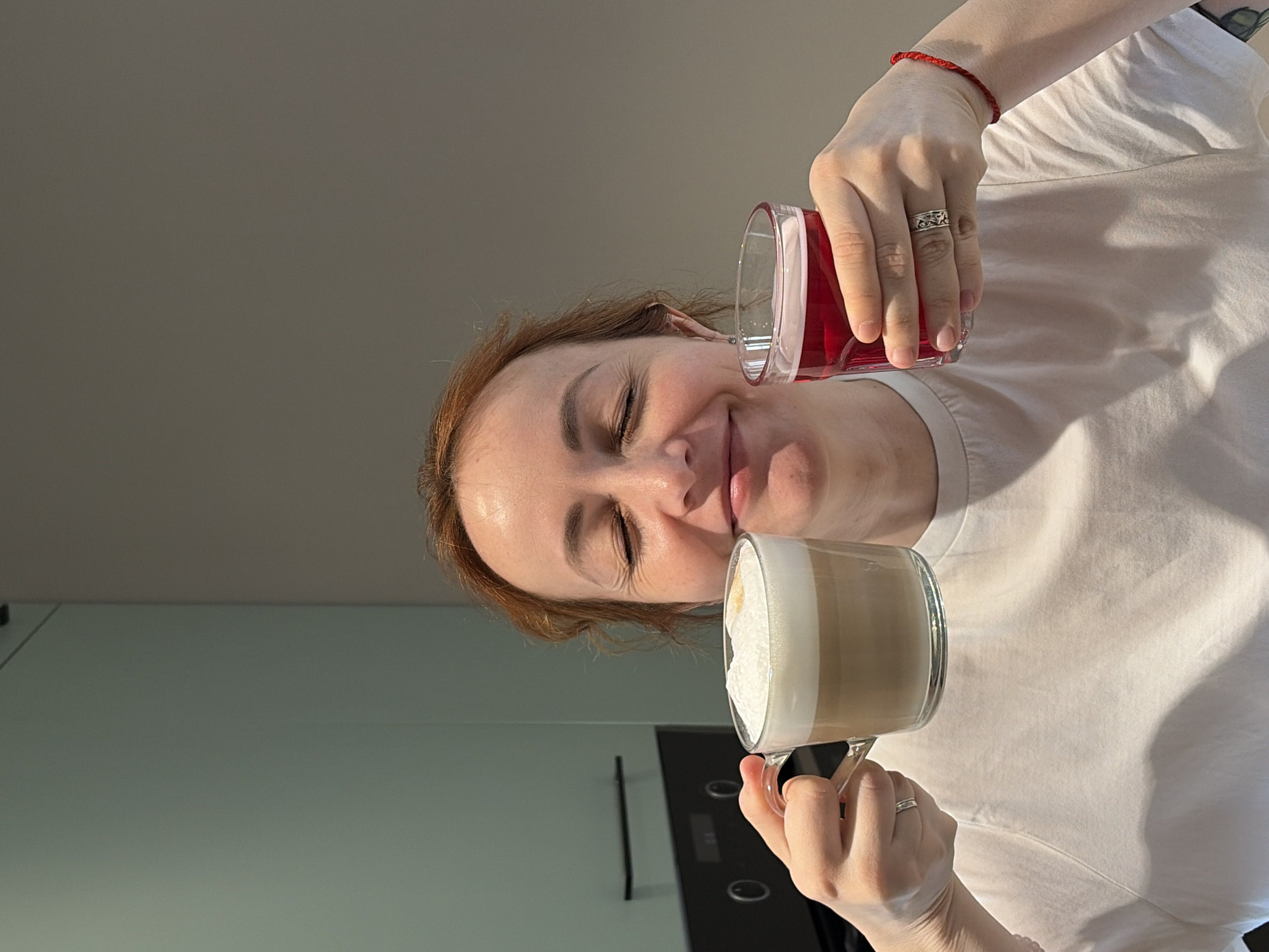 A woman is holding a glass of red liquid over a glass of coffee in what appears to be a kitchen. She is wearing a white t-shirt and has a red bracelet on her wrist. She has a ring on her finger. The kitchen has light green walls and black appliances. There is no explicit information about the location being in Chaikovsky, Russia.