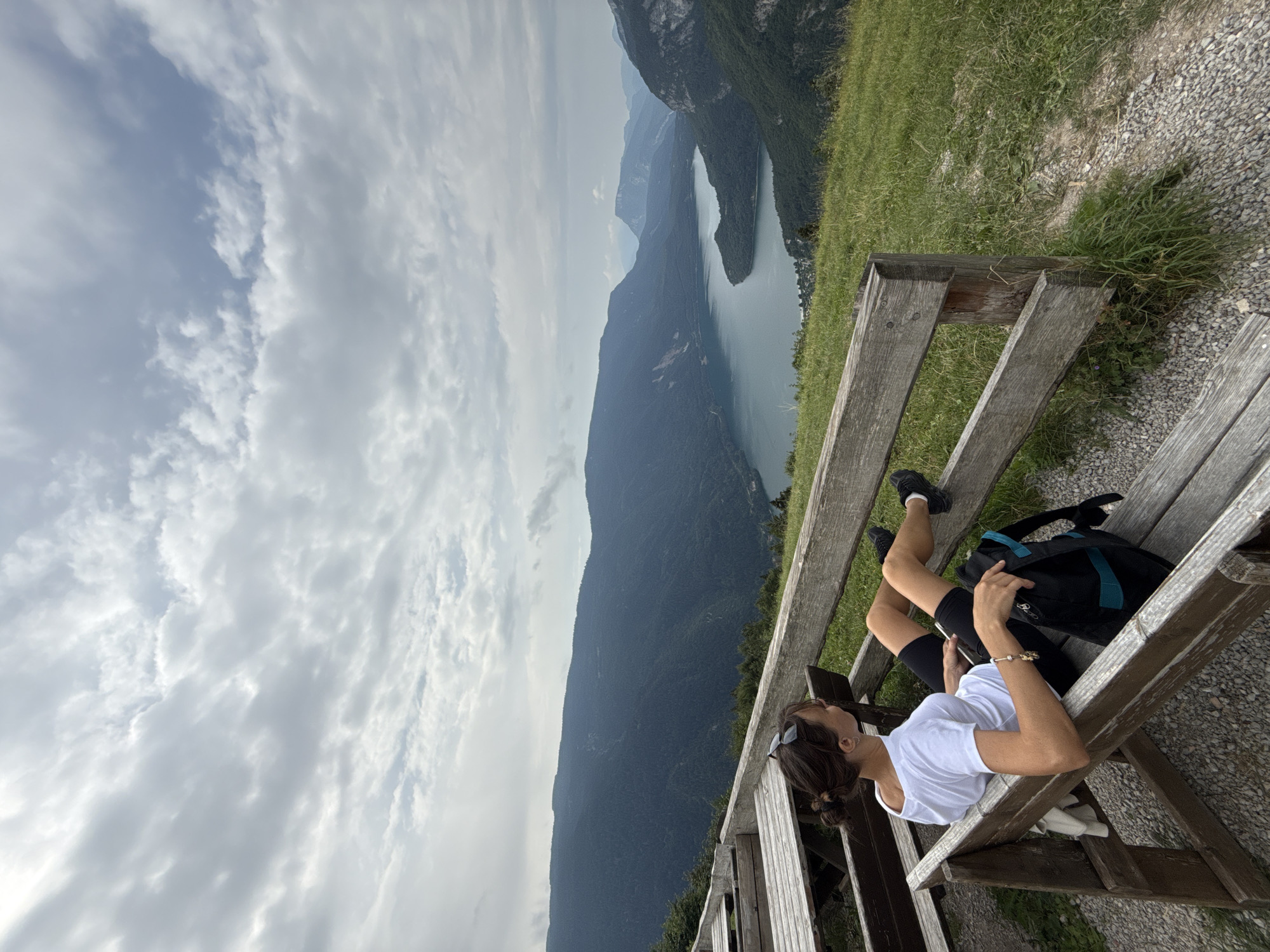A woman is sitting on a wooden bench with her legs extended, looking out at a panoramic view of a lake surrounded by mountains. The woman has her hair pulled back and is wearing a white t-shirt and black shorts. A backpack is on the bench next to her. The scene is outdoors, with a cloudy sky overhead. The location is the Molveno Overlook in Molveno, Italy, offering a scenic view of Lake Molveno and the surrounding Dolomite mountains.