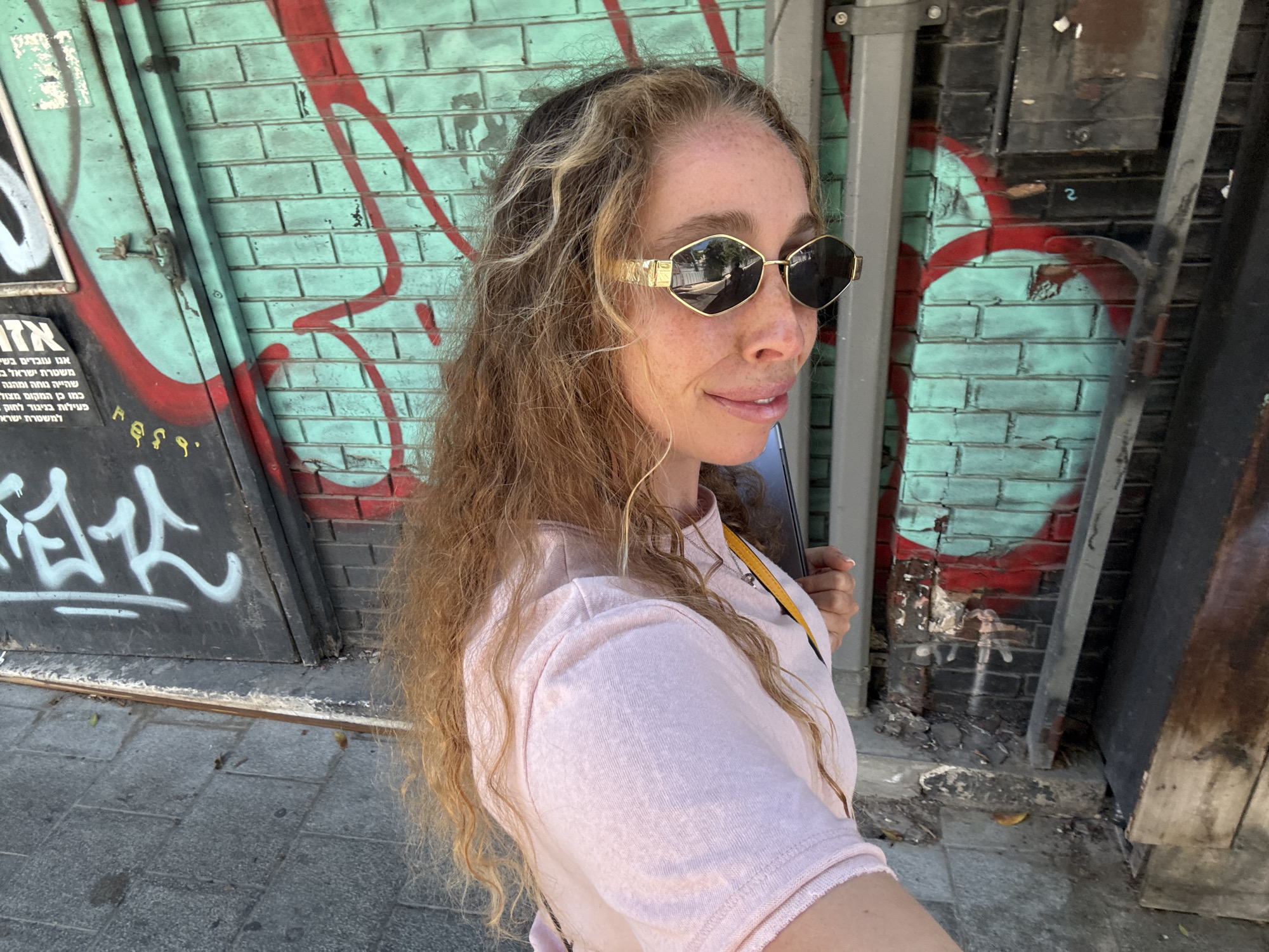 A woman with long, wavy blonde hair and freckled skin is captured in a close-up, side-facing shot. She is wearing a light pink t-shirt and distinctive gold-rimmed sunglasses with a hexagonal shape. Her expression is a gentle smile. She is holding a dark object, possibly a phone or tablet, in her right hand.
The background features a brick wall covered in graffiti. The bricks are painted a light teal color, with bold red spray paint markings in a swirling pattern. Part of a metal door or structure with graffiti is visible to the left, displaying Hebrew text. The ground is paved with gray stones. The lighting suggests it is daytime, possibly late morning or early afternoon, with bright sunlight casting shadows. The scene appears to be an urban environment, with the graffiti and weathered brick suggesting an outdoor, possibly slightly gritty, location. The visible Hebrew text, "אנו עובדים בשם משטרת ישראל" which translates to "We are working in the name of the Israel Police", and other phrases, along with the location context of Tel-Aviv, Israel, confirms the setting. The overall mood is casual and candid.