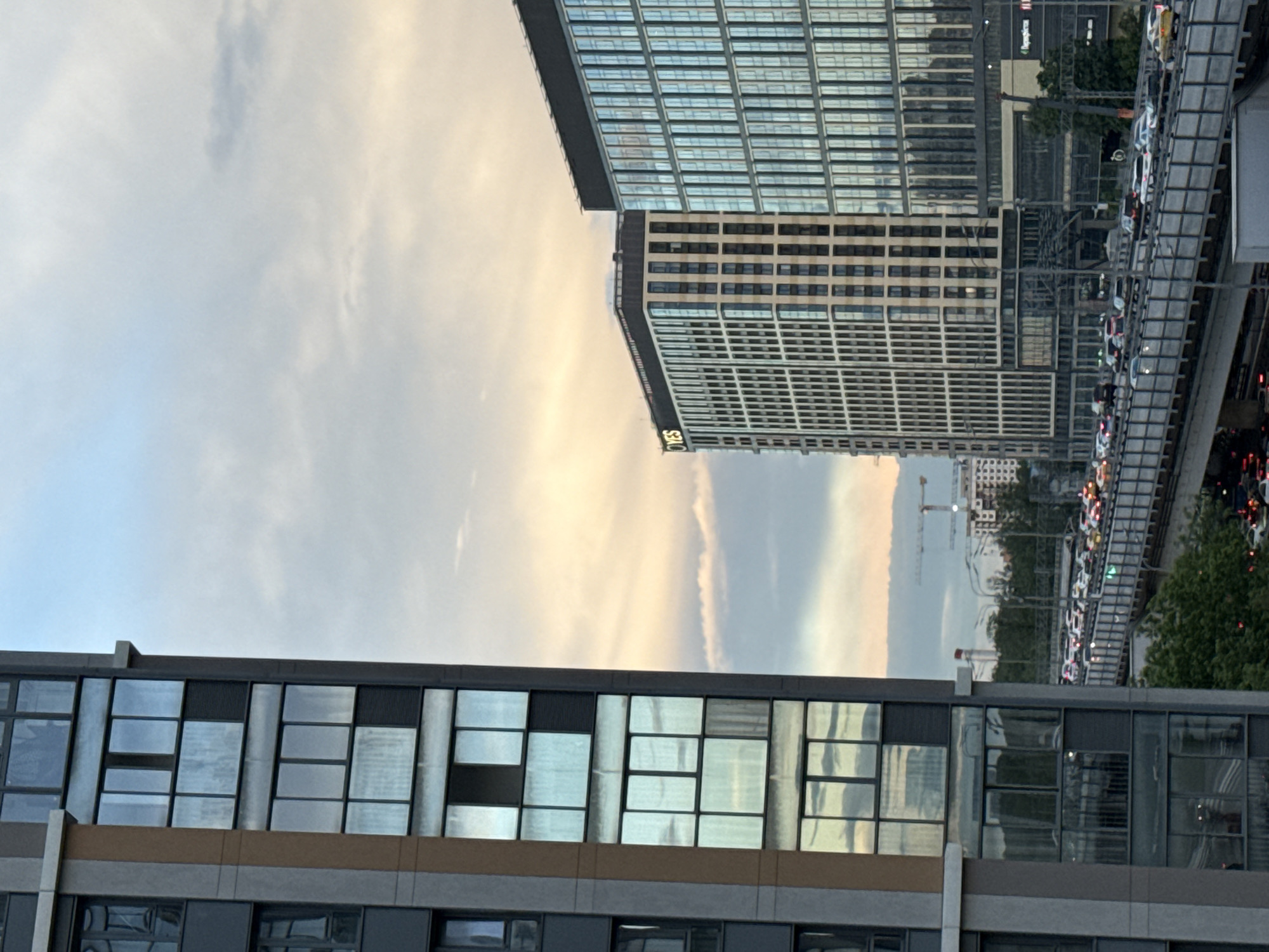 The image shows a cityscape at dusk or dawn, with a modern building featuring large, reflective windows in the foreground and a taller, grid-patterned skyscraper in the background. A multi-lane highway overpass runs adjacent to the skyscraper, filled with traffic. A crane is visible in the distance behind the highway, suggesting ongoing construction. Given the location identified as Khimki, Russia, it is likely an urban area with active development and transportation infrastructure. The sky has a mix of clouds with the yellow hue of sunset or sunrise.