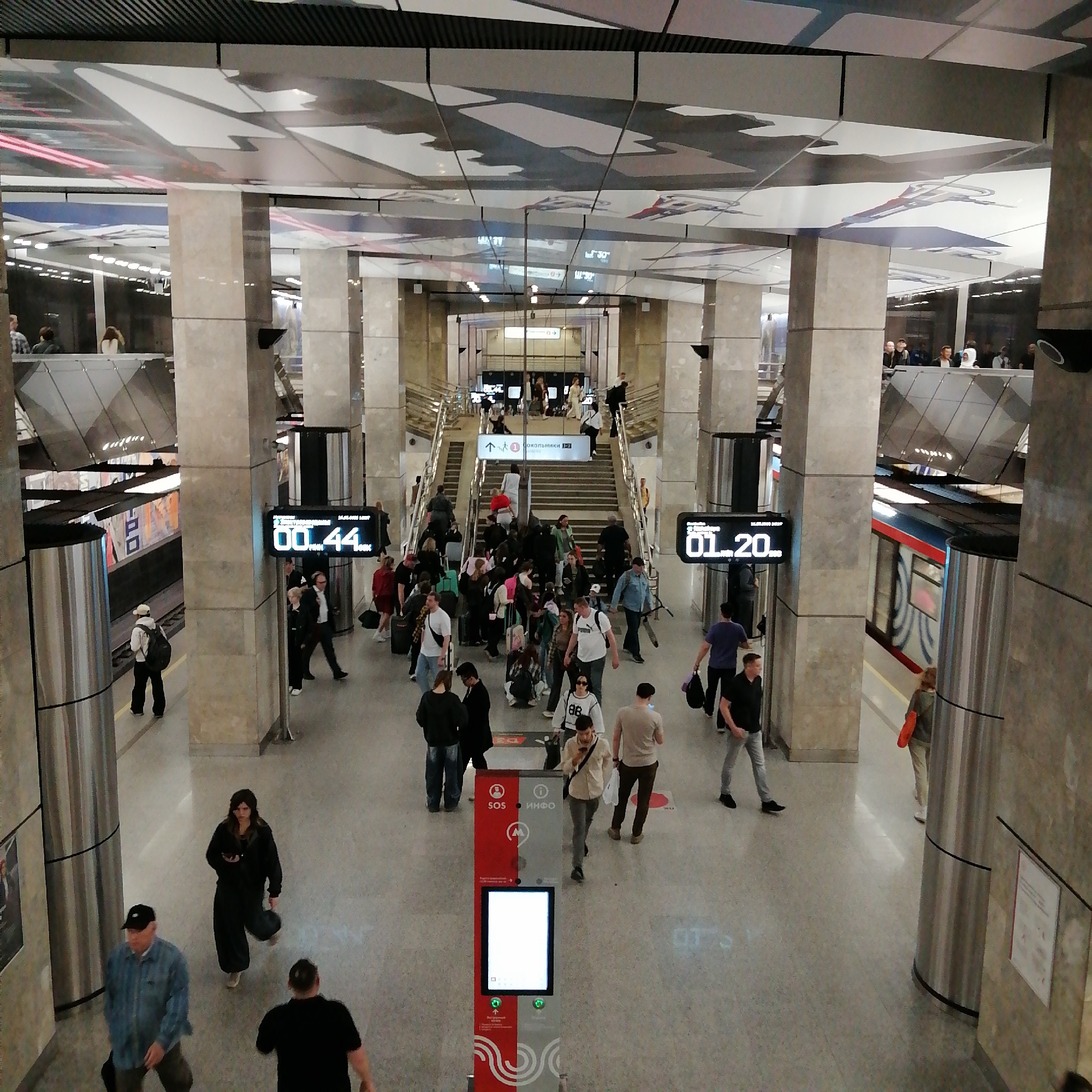 The image depicts the interior of a Moscow Metro station, likely in Russia. The scene is a busy intersection with people moving in various directions. Many individuals are traveling up and down stairs leading to a higher level, while others are walking across the main floor. Several people are carrying luggage or bags, suggesting they may be commuters or tourists. Electronic displays on columns show train arrival times. A train is visible on the right platform. A red and gray information kiosk with a digital screen is prominently positioned in the foreground. The station's architectural design features large columns and a decorated ceiling with what appears to be artwork. The color palette of the station primarily comprises neutral tones like gray, beige, and white, with accents of red and blue from the trains and signage.