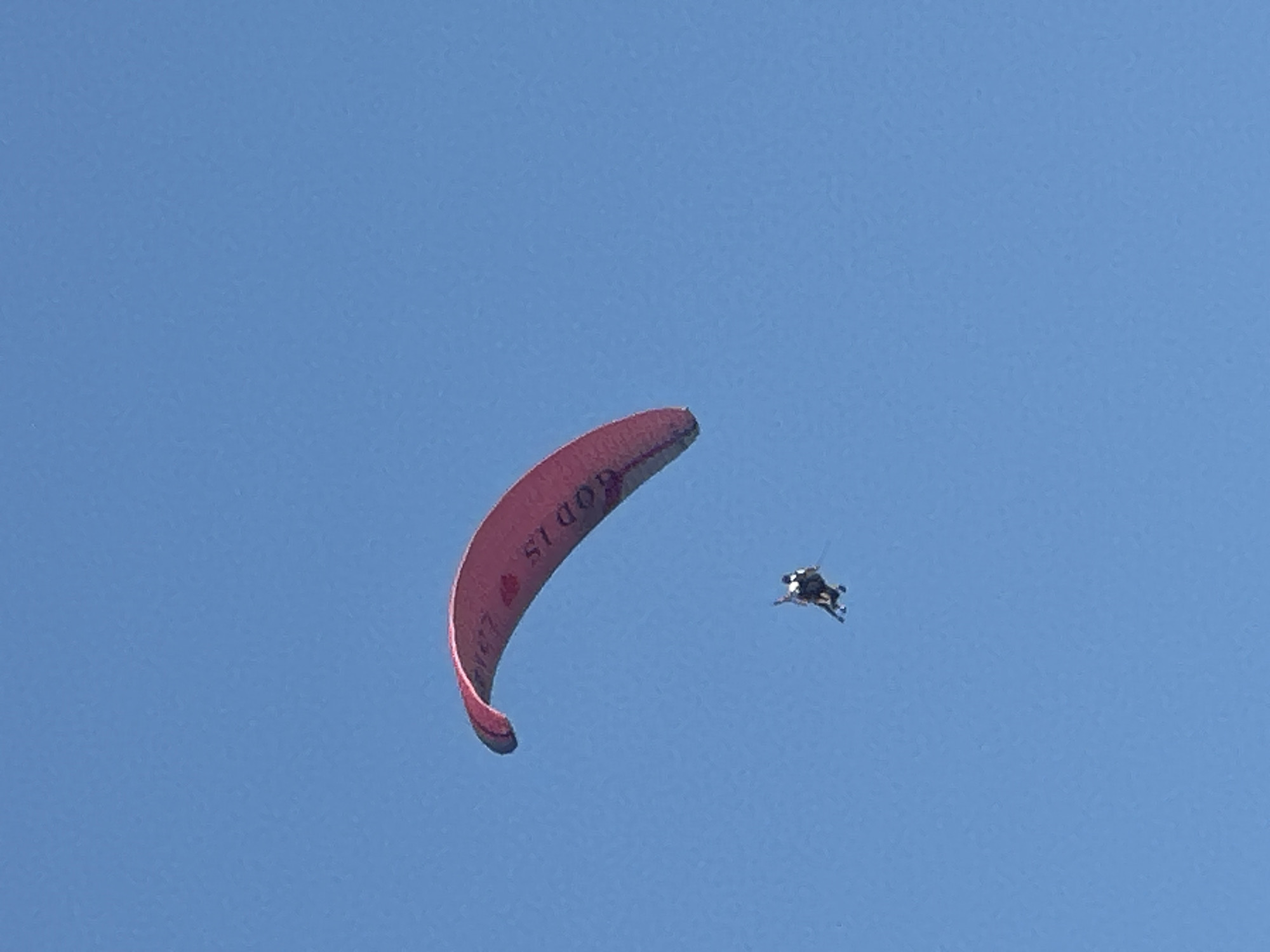 A tandem paraglider is captured mid-flight against a clear blue sky. The paraglider's canopy is a vibrant red with dark text that appears to read "GOD IS LOVE". Below the canopy, two individuals are strapped together, appearing to be the pilot and passenger, suspended in the air. The paraglider is positioned in the upper left quadrant of the frame, with the people directly beneath it and slightly to the right. The image offers no discernible details about the city of Boreti, Montenegro.