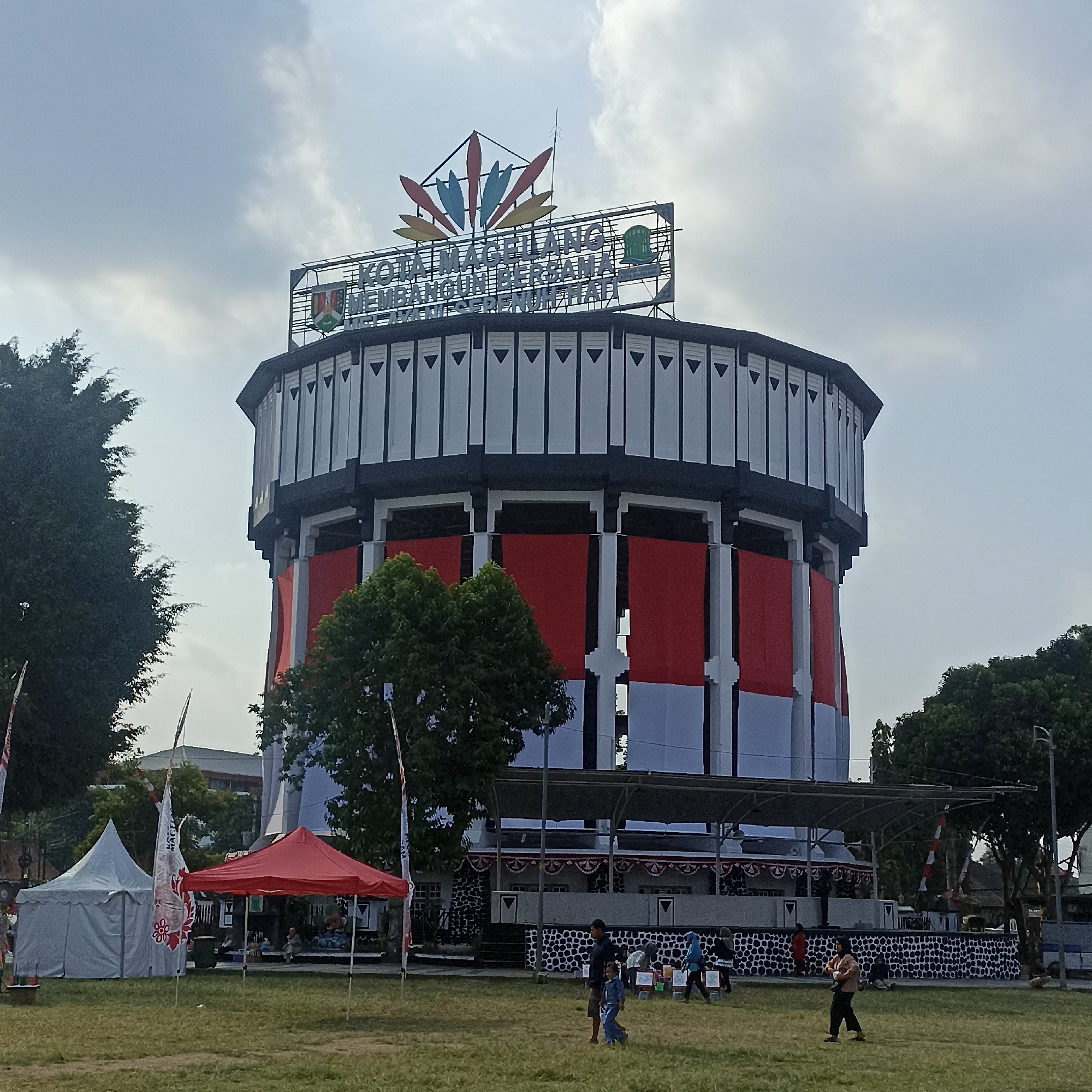The image captures an exterior view of the Grand Magelang building, located in Magelang, Indonesia. The structure is a large, cylindrical building with a distinctive tiered roof. The facade is adorned with alternating red and white banners, evoking the colors of the Indonesian flag. The building is topped with a prominent sign displaying the words "KOTA MAGELANG MEMBANGUN BERSIH DAN SEHAT."
In the foreground, a grassy field hosts several tents, including a white dome tent and a red canopy tent. A few individuals are present on the field, some walking and others sitting. The sky above is overcast with scattered clouds. Trees are visible on the left and right sides of the image, framing the central building. The overall scene suggests an outdoor event or gathering.