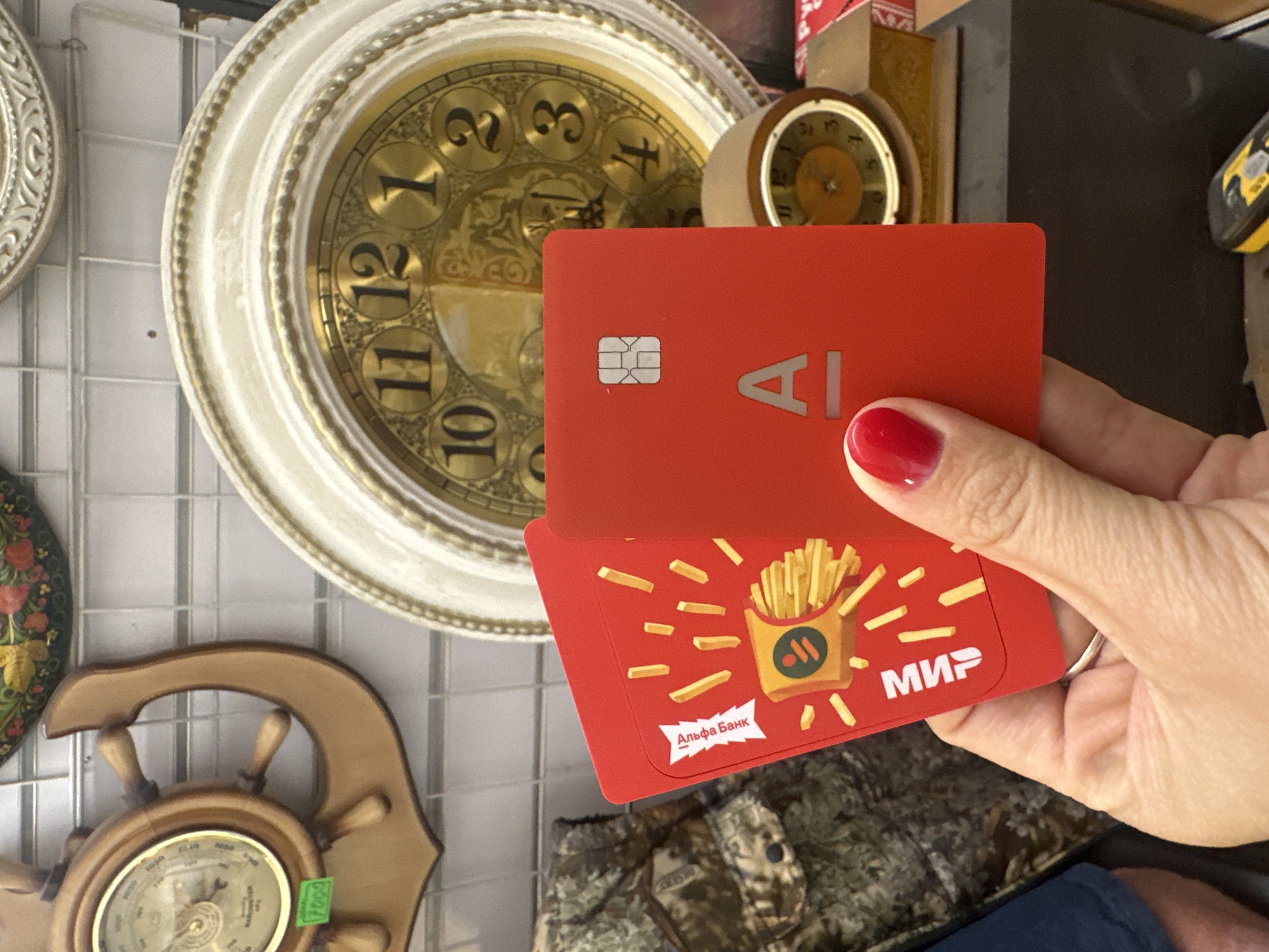 In Novoaltaysk, Russia, a person's hand with red nail polish is holding two red cards. The card on the left displays an illustration of a french fry container and the text "Альфа Банк" and "МИР". The card on the right has a chip, the letter "A", and a horizontal line. In the background, a large circular clock with Roman numerals is visible, along with a ship's wheel barometer and other decorative items.