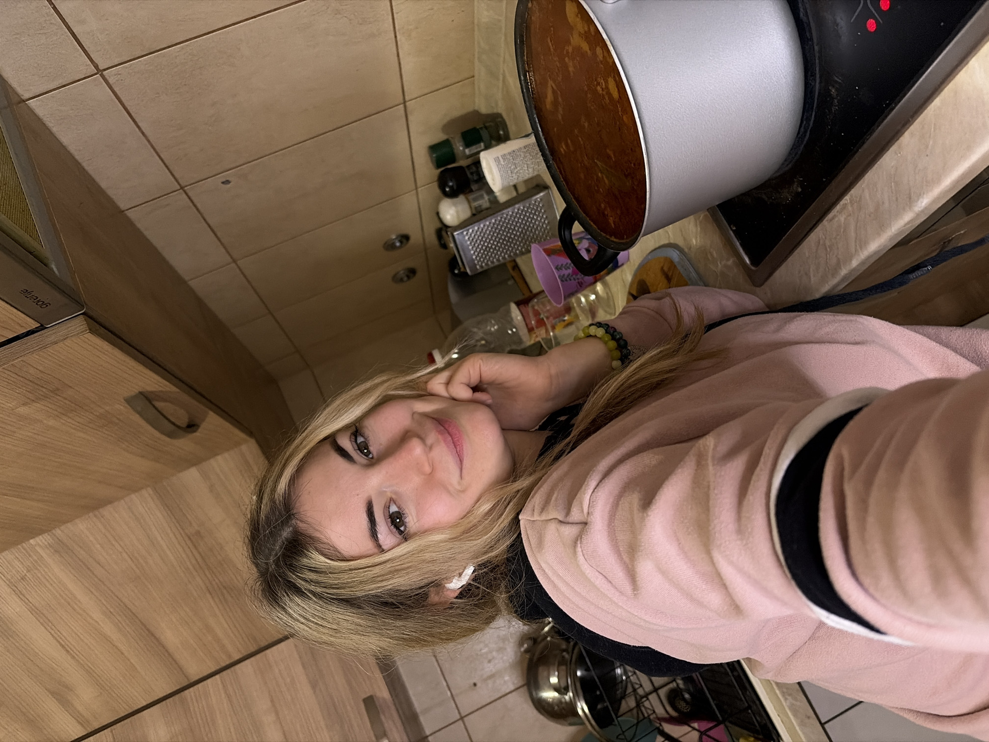 A woman is in a kitchen, possibly in Budva, Montenegro. She has blonde hair and is wearing a pink sweater. In the background, a pot of soup sits on a stove. There are various kitchen items visible, including spices and a grater.