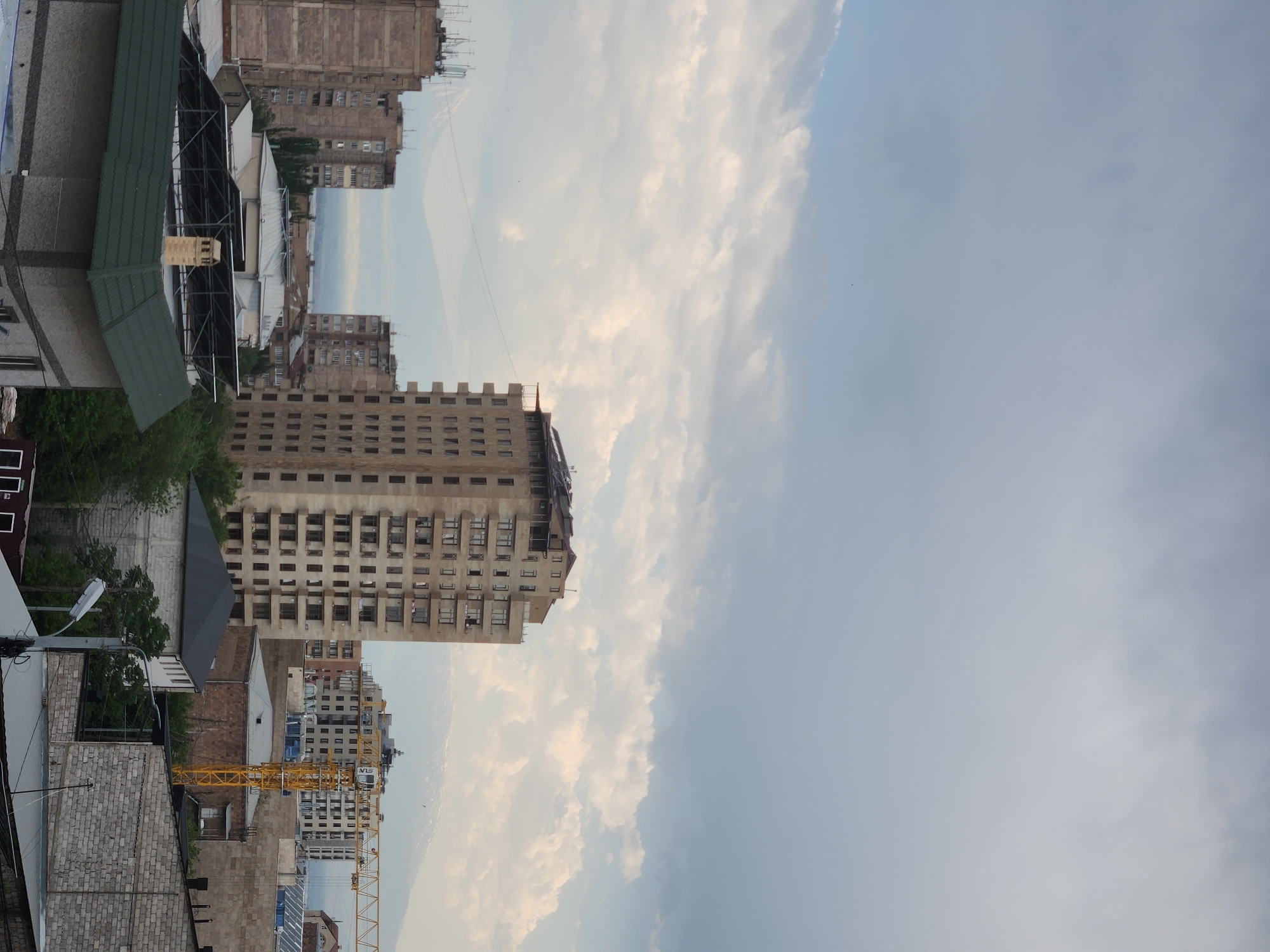 This is an aerial, eye-level, medium shot of a city skyline under a cloudy sky. The main focus is a tall, unfinished apartment building in the center of the frame. It is made of concrete and has many windows. To the left of this building are two smaller apartment buildings, one of which has antennas on top. Further in the background, snow-capped mountains are visible. To the right of the unfinished apartment building, another apartment building and a yellow construction crane can be seen. In the foreground, there are several rooftops, including one with solar panels and a chimney. The environment appears to be urban, with a mixture of residential and commercial buildings.