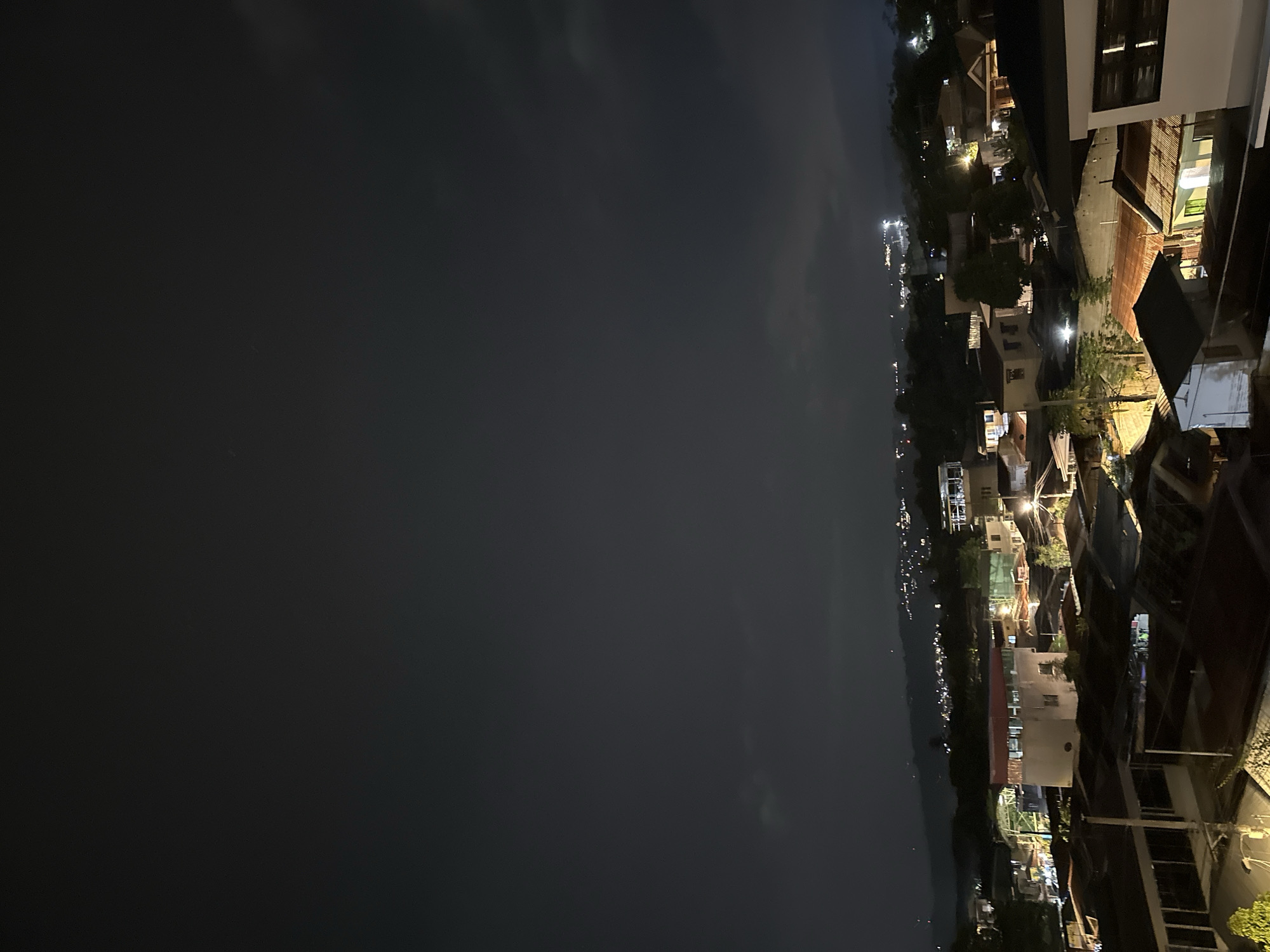 The image shows an urban landscape at night, likely taken from an elevated position in Davao City, Philippines. The right side of the frame is filled with buildings and houses, varying in size and architecture. Artificial light illuminates these structures, creating pools of brightness against the dark. Streetlights are visible along the winding road, further illuminating the scene. The left side of the image is occupied by a dark expanse, possibly a body of water or a large open space, reflecting minimal light. The overall scene is a mix of densely built-up areas and dark, open spaces.