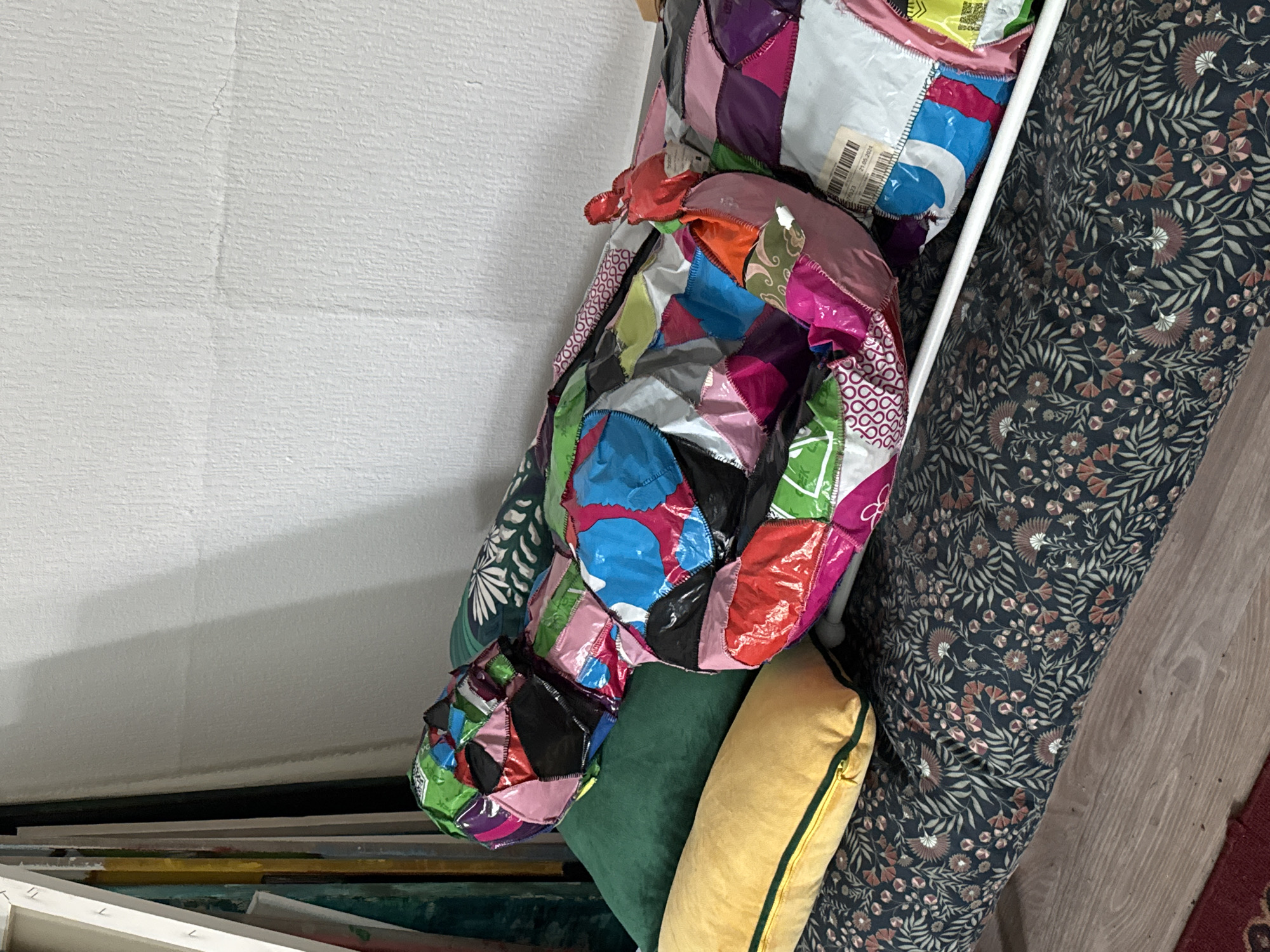 The image shows a life-sized mannequin covered in a patchwork of colorful, shiny material, resting on a bed of pillows and a patterned mattress. The mannequin is positioned as if lying down, with its head and torso clearly visible. The patchwork material is made up of squares and rectangles of various colors including pink, green, blue, black, red, and purple. Some of these pieces appear to be made from recycled packaging materials, as evidenced by printed text and labels visible on some sections.
The mannequin is lying on top of several pillows: a large, dark green pillow is positioned behind the mannequin's head and torso, and a long, yellow pillow is placed in front of it. Beneath the pillows and mannequin is a mattress with a dark fabric covering featuring a repeating floral and leaf pattern in muted tones of blue, green, and rust.
To the left of the scene, several canvases or framed artworks are propped up against a white support structure. The background is a plain, textured white wall, with a faint horizontal line suggesting a seam or crack in the plaster. The flooring visible at the bottom of the image is light-colored wood.
The image does not provide any explicit information about the city of Moscow, Russia, or any specific location within it.