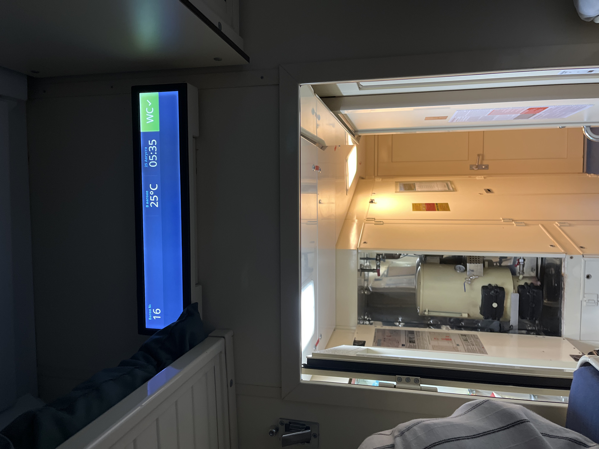 The image captures an interior view of a train compartment, focusing on a digital display screen mounted above an open doorway. The screen shows information including the wagon number (16), the date (August 25th), the time (05:35), the temperature (25°C), and the status of the WC (indicated by a green checkmark). To the left of the screen, a blue blanket is visible, draped over a bunk. The open doorway reveals a corridor with a brightly lit interior. Inside the compartment to the right of the doorway, there is visible mechanical equipment. The overall environment appears to be clean and functional, consistent with a train carriage. The location is identified as a train in the city of Krugloye, Russia.