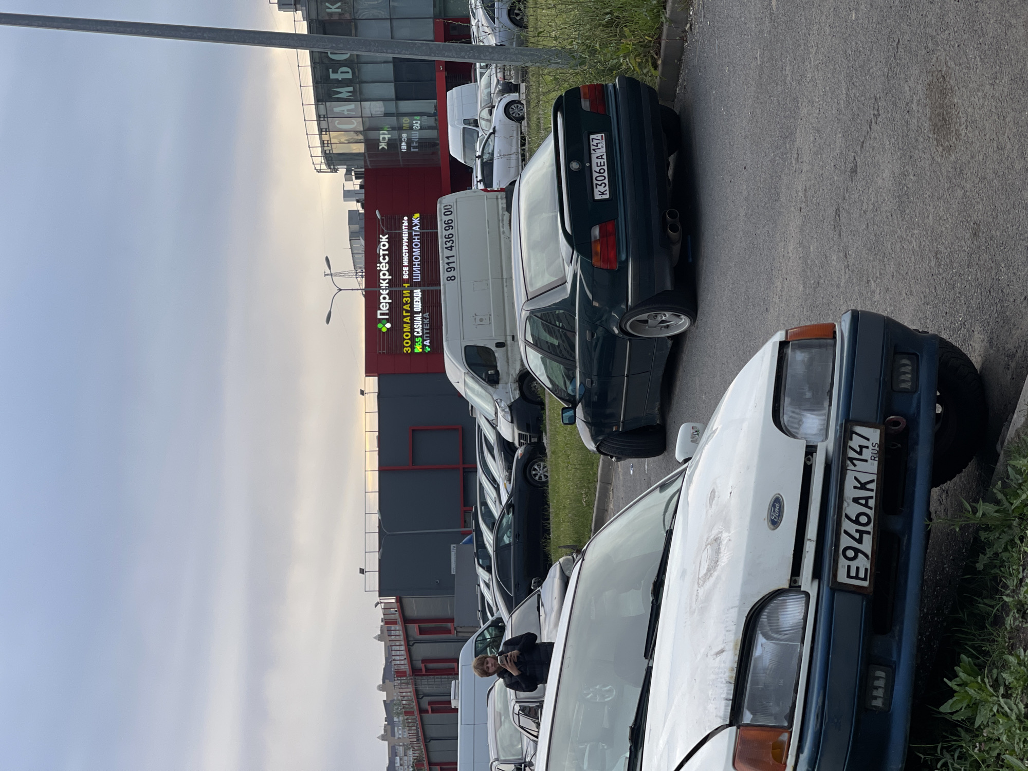 A medium shot captures a parking lot scene with several cars, a building, and a person. In the foreground, the hood and front bumper of a white Ford are visible, displaying a license plate that reads "E946AK 147 RUS". To the right of the Ford, a dark green BMW sedan is parked, its rear facing the camera. Its license plate shows "K306EA 147 RUS". In the background, a large building with red and dark grey sections is prominent. A large illuminated sign displays "Перекрёсток" in Russian, along with other text and logos. To the right of this building, another building with large glass windows and white lettering that appears to read "САМБО" is visible. A person with light brown hair, wearing a dark jacket, stands between parked cars, holding a phone and appearing to take a photo or video. The sky is overcast with clouds. The environment is an outdoor urban setting, likely a commercial area or shopping center parking lot.