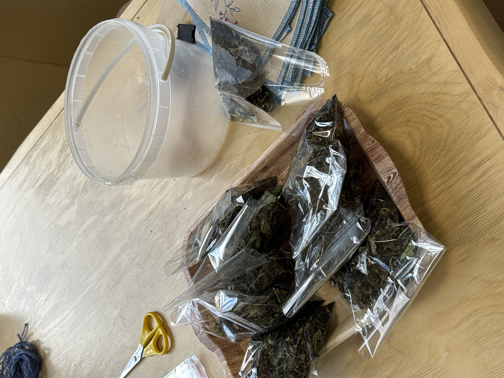 The image shows a wooden table with several items on it. In the foreground, a wooden tray holds multiple small, clear plastic bags filled with dried green leaves. To the left of the tray are a pair of yellow-handled scissors and a ball of dark blue yarn. In the background, to the right, is a clear plastic bucket with a light-colored handle. A stack of folded cloths with blue trim is placed next to the bucket. To the right of the tray, a clear plastic cup, partially filled with dried leaves, is also visible. The scene is lit from above, casting subtle shadows on the table surface. The overall impression is one of preparation or organization of dried plant material. The context of Gora (Khokhlovskoe S/P), Russia is not directly observable from the image itself.