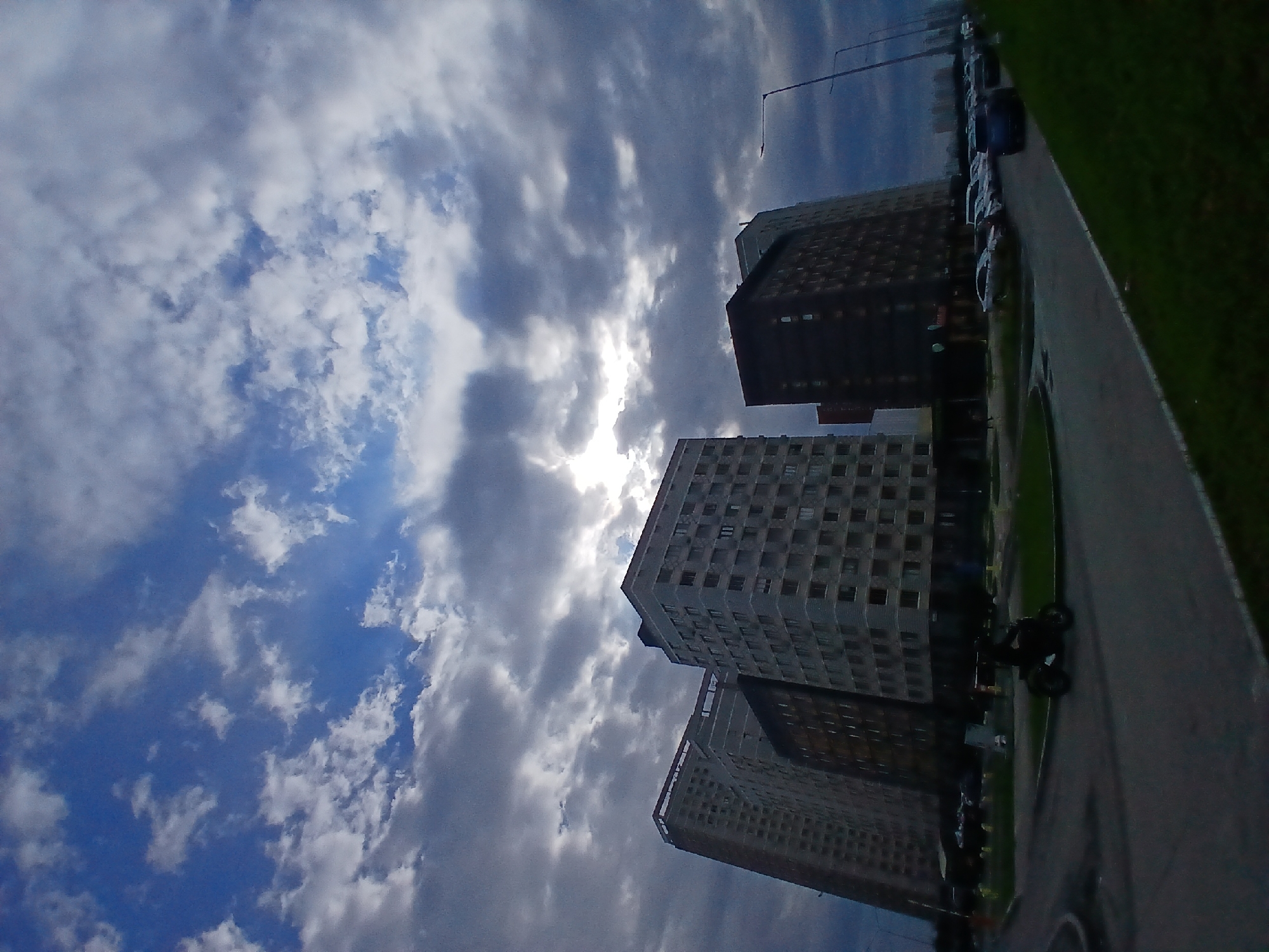 The image shows a street scene in Yekaterinburg, Russia, featuring several tall apartment buildings under a cloudy sky. The buildings are modern in construction, with visible windows and balconies. In the foreground, a dark-colored motorcycle with a rider is moving on a paved road. To the right of the road, a row of parked cars is visible, and beyond that, a patch of green grass. Streetlights are also present along the road. The sky is dramatic with a mix of dark and light clouds, with the sun partially visible through a break in the clouds.