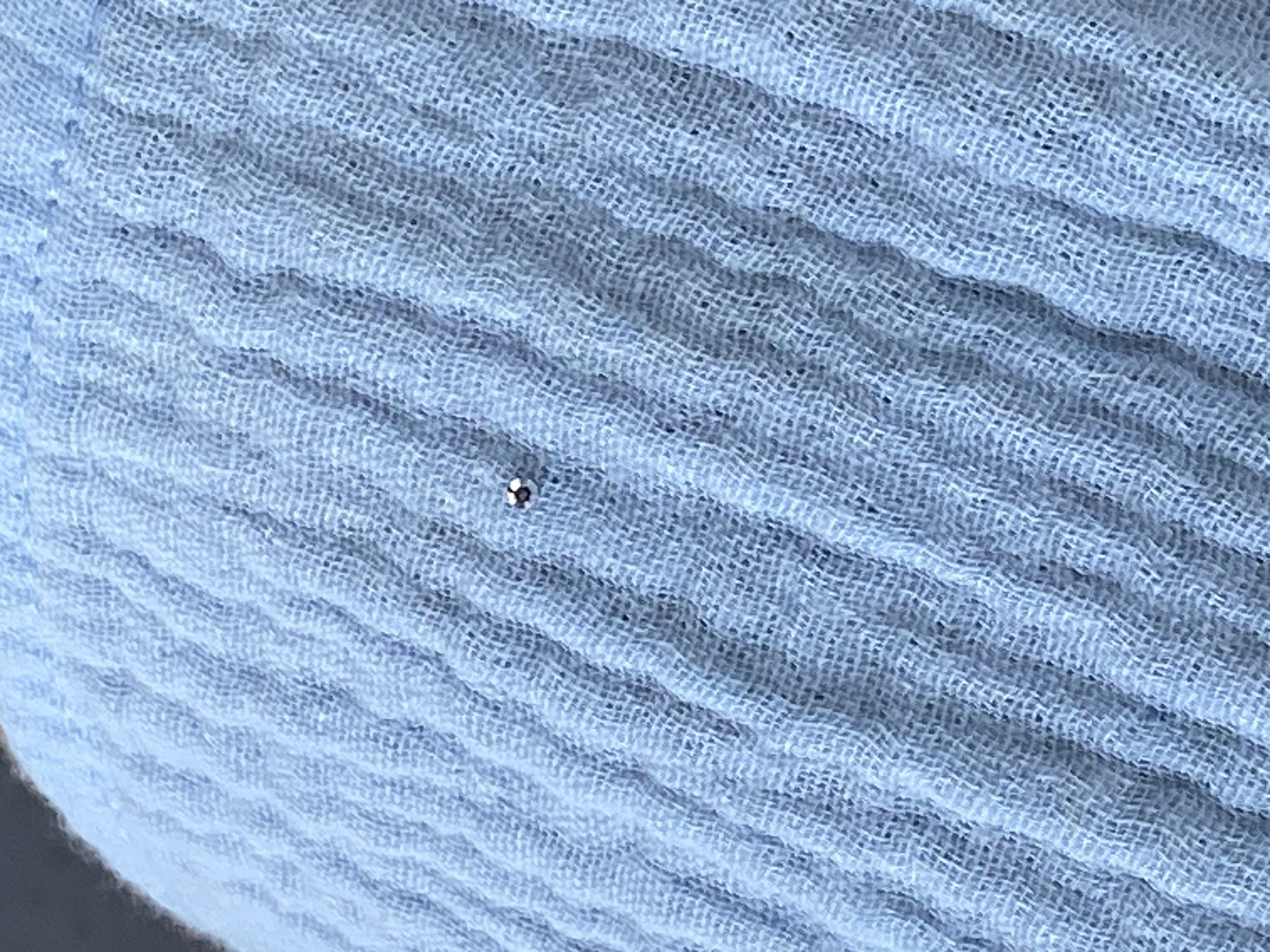 A close-up, eye-level shot shows a small, metallic bead or charm resting on a textured, light blue fabric. The fabric appears to be a lightweight material with a crinkled or wrinkled surface, possibly muslin or a similar textile. The bead is positioned slightly off-center to the right of the frame. The background is out of focus, but the texture of the fabric dominates the image. There is no information about the location in the city of Bac Nha Trang Ward, Vietnam.