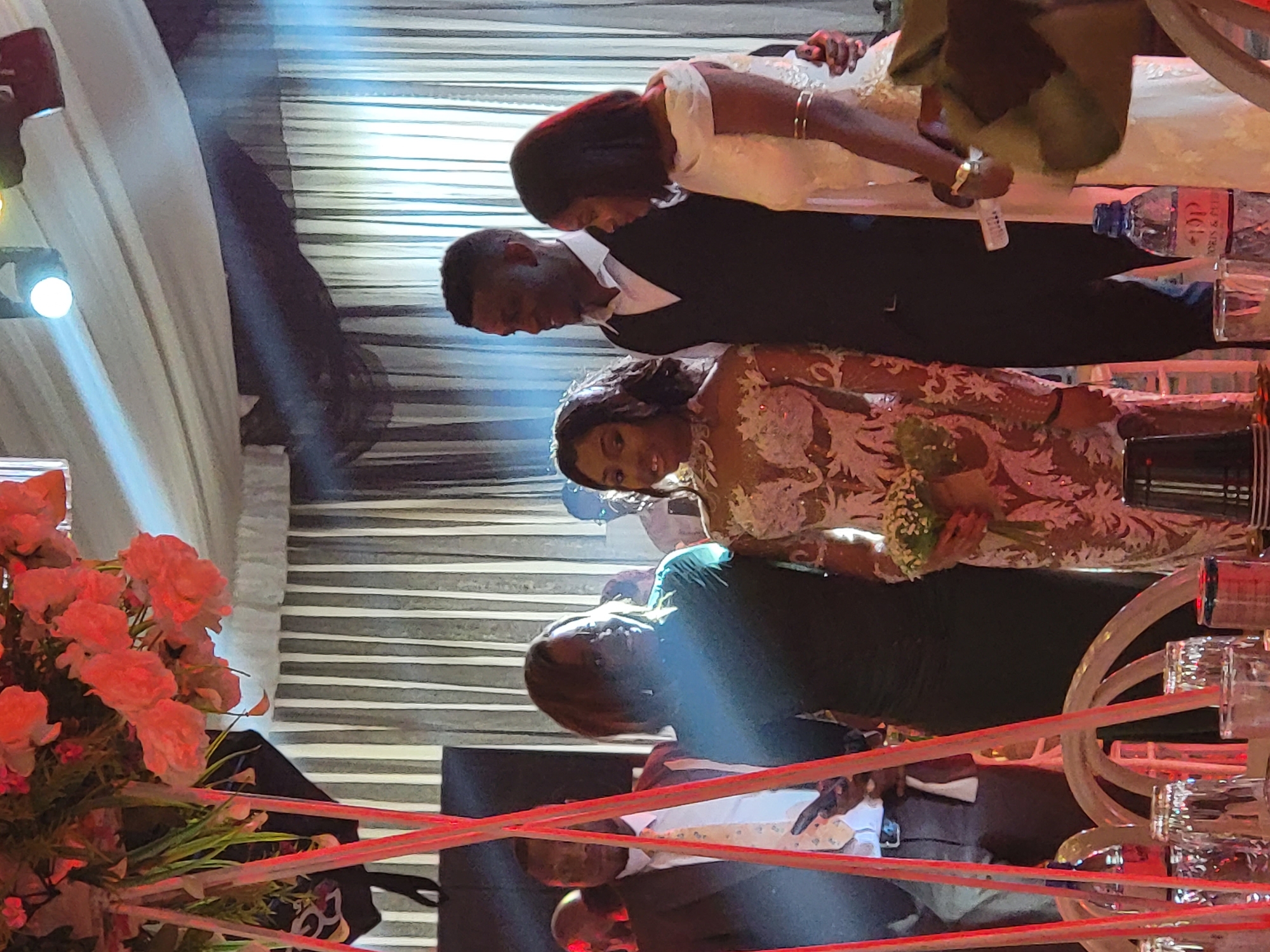 An indoor formal event, possibly a wedding reception, is captured with a celebratory atmosphere under artificial lighting, suggesting an evening gathering in Barden Yana, Nigeria.
In the center-back, a man in a black suit and white shirt stands beside a woman in a white off-shoulder gown, likely the groom and bride, both looking downwards. In front of them, a woman in an elaborate, possibly lace, dress holds a small white bouquet and looks directly towards the viewer. Another woman in a dark dress is partially visible behind her. In the lower foreground, a man in a white shirt and dark jacket is also present.
The scene includes a vibrant bouquet of pink and red flowers positioned in the far left foreground. To the right, a water bottle with the brand "FIJI" partially visible on its label, along with several glasses, are placed on a surface. Red decorative cords or lighting elements span across the bottom of the frame, creating a foreground barrier. The background features white drapes or curtains and walls with horizontal striped patterns. Bright stage lights and spotlights illuminate the area, causing some lens flare in the image.