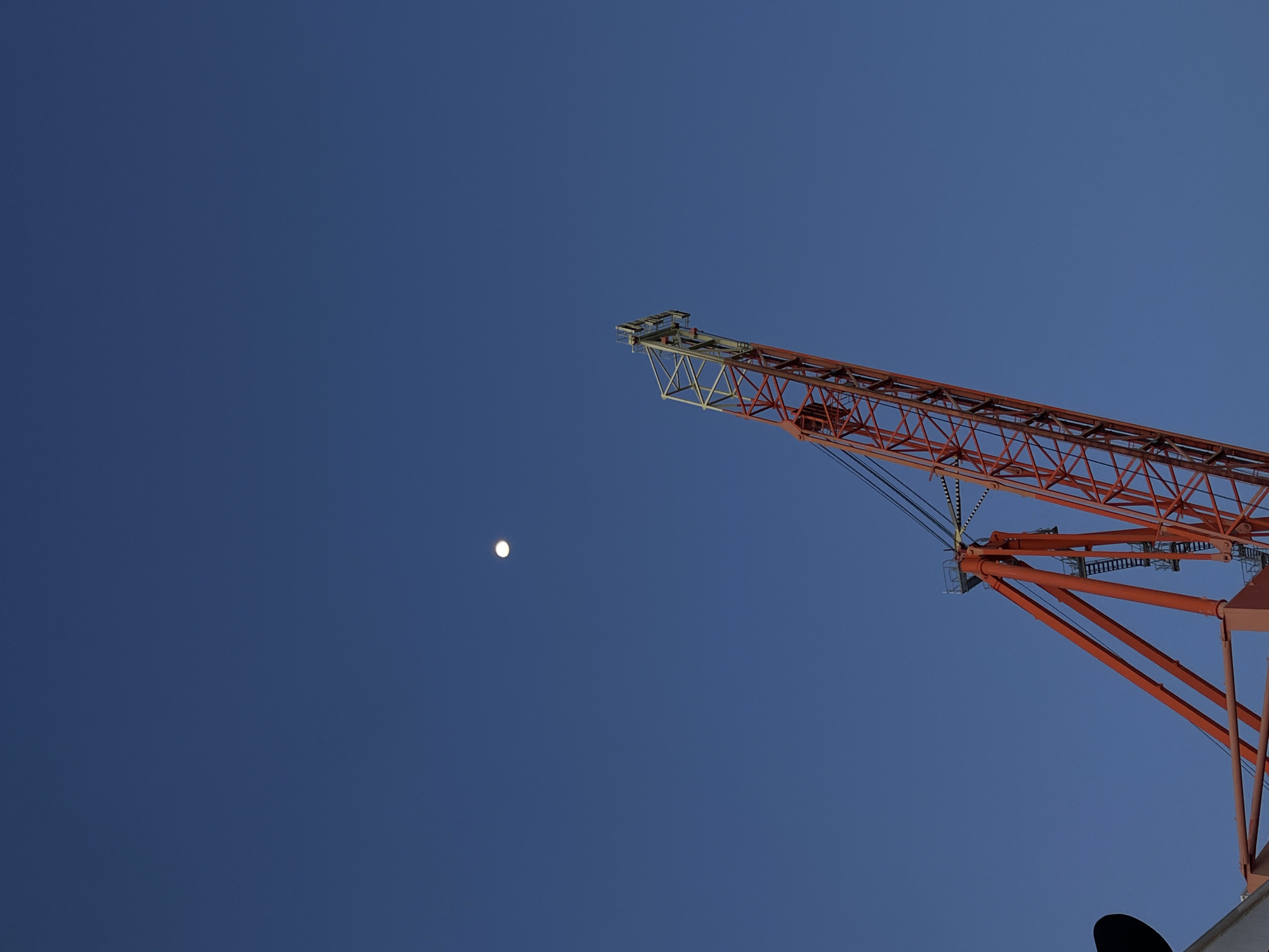 The image captures a striking view of a large, red-orange industrial crane set against a vast, clear, deep blue sky. The crane's intricate lattice-like arm extends diagonally from the lower right corner of the frame, reaching upwards and across the mid-right section. Its metallic structure, comprised of numerous beams and supports, is clearly visible, along with some connecting cables near its main joint.
In the upper-left portion of the sky, a small, bright white circular object is prominently featured, which is identifiable as the moon. The crisp visibility of both the moon and the consistent deep blue of the sky suggests a clear day, likely either morning or late afternoon, as the moon can be seen during daylight hours under such conditions.
There are no people or discernible activities taking place in the scene, which appears static and focused on the architectural presence of the crane against the natural backdrop of the sky. No visible text can be found within the image. The setting is implied to be an outdoor industrial or construction environment, given the presence of such a large-scale crane. The overall mood is one of stark minimalism, highlighting the contrast between the man-made structure and the celestial body.