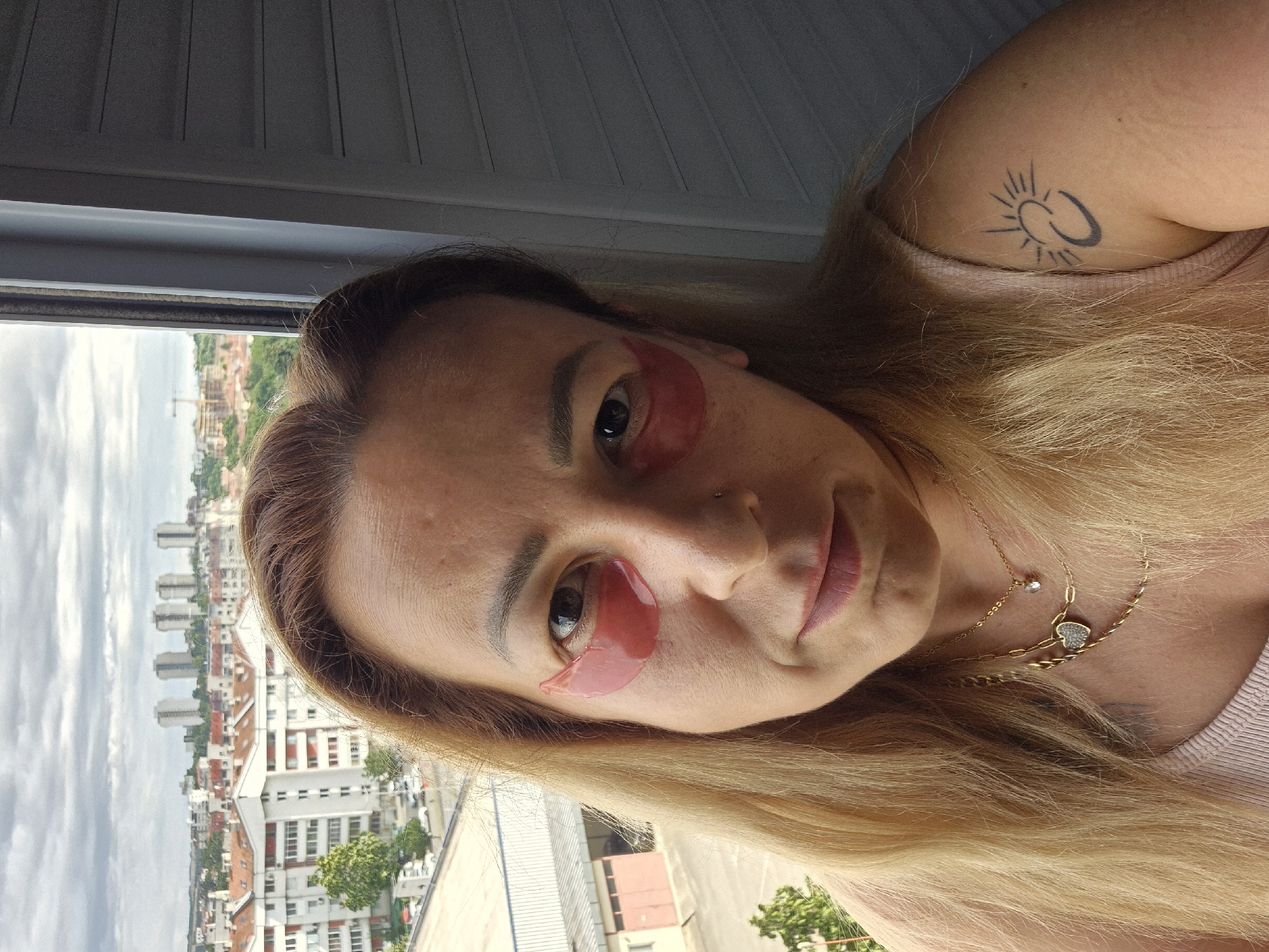 In an urban setting, a woman with blonde hair and eye patches is captured in a close-up, self-taken photograph. The background reveals a cityscape with buildings and a cloudy sky. She is wearing necklaces and has a tattoo on her upper arm. The lighting suggests it is daytime, and the focus is on her face and upper body.