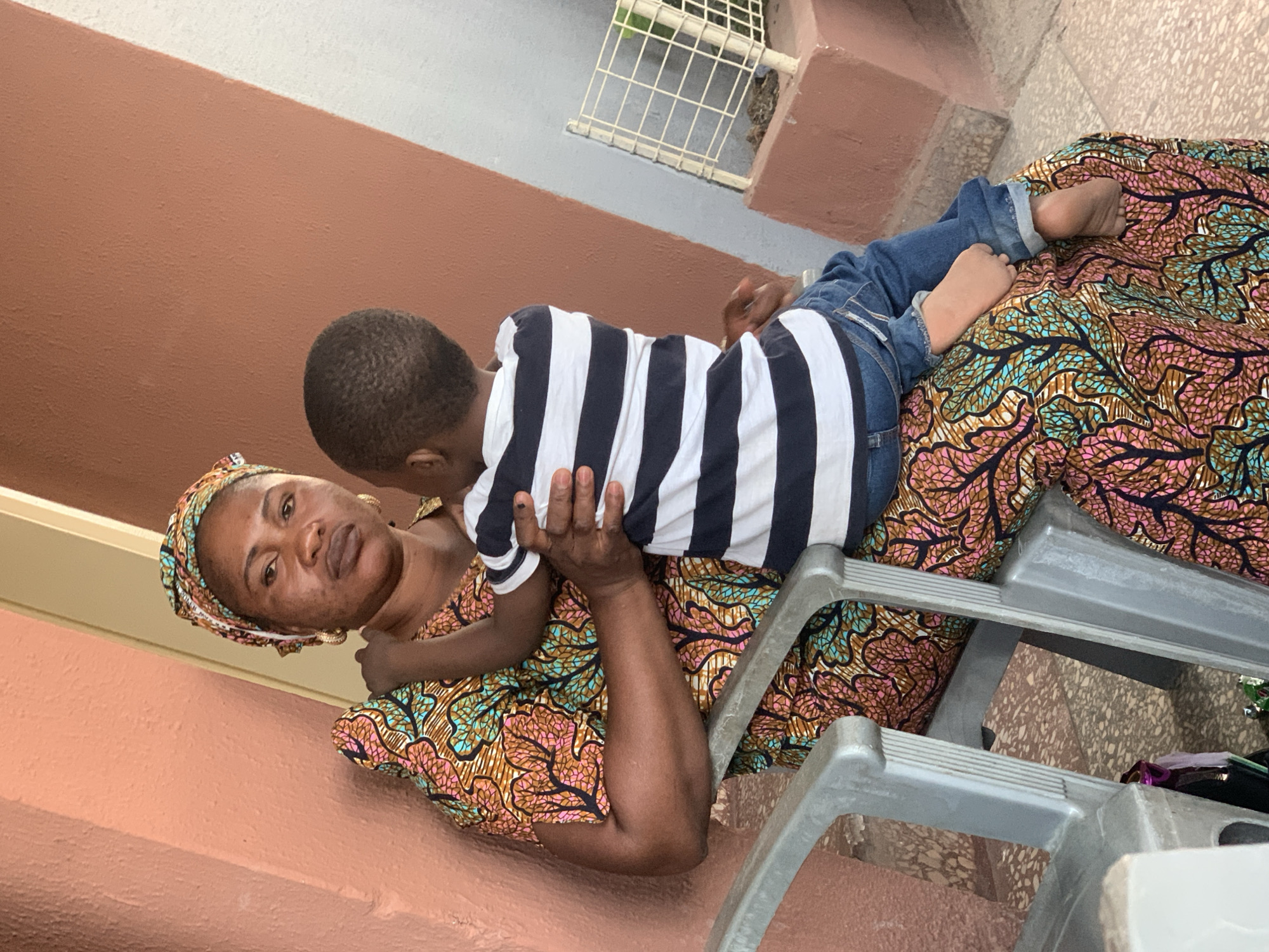 A woman and a child are on an exterior surface in Jos, Nigeria. The woman is lying on her back, wearing a colorful patterned headwrap and a dress with similar patterns. She is holding a young child who is lying on top of her. The child wears a black and white striped shirt and blue jeans. The woman's patterned dress partially covers what appears to be a gray plastic step stool. The background consists of a brown and gray wall, and part of a white barred structure is also visible. The lighting suggests it is daytime.