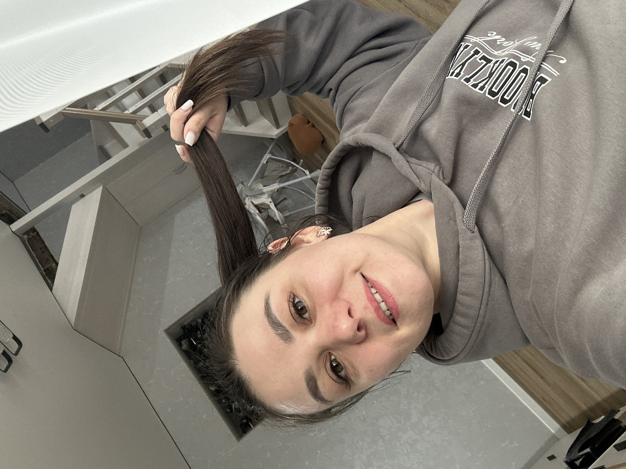 The image captures a young woman posing for a selfie-style photograph, rotated clockwise. She is the central subject, holding a long, thick strand of her dark brown hair with her right hand, which features white manicured nails. She looks directly at the camera with a slight, pleasant smile, showcasing light pink lipstick. Her eyes are brown, and she has well-defined eyebrows. A small, ornate earring is visible in her right ear.
She is wearing a grey hooded sweatshirt. On the front of the sweatshirt, the word "BROOKLYN" is clearly visible in a collegiate-style font, with "New York" written above it in a cursive script.
The background suggests a modern residential interior in Chaikovsky, Russia. In the upper left, a staircase with light-colored risers and wooden treads can be seen. Further into the background, a white baby swing or playpen is partially visible, next to a brown, soft object, possibly a cushion. The flooring appears to be a mix of light-colored surfaces and wooden sections. The lighting indicates an indoor setting, likely during the daytime or in a well-lit space. The image conveys a casual, everyday moment.