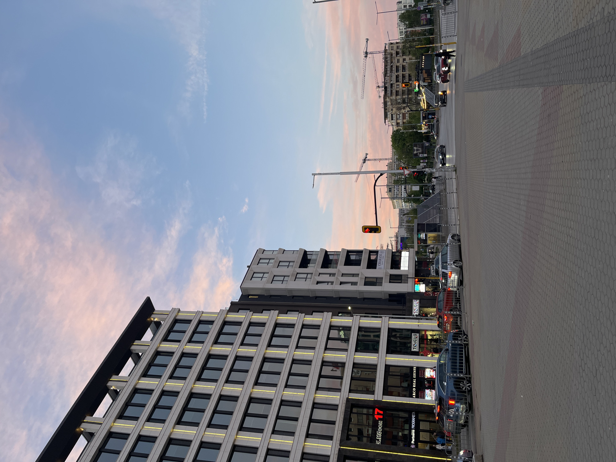 The image depicts a street scene in Sofia, Bulgaria at dusk. The sky is a mix of blue and pink with light clouds. In the foreground, there's a modern building with a grid-like facade and visible lights. Adjacent to it is another building with shops at street level. A traffic light shows a red signal. Cars are parked along the street, and pedestrians are seen on the sidewalk. Construction cranes are visible in the background, suggesting ongoing development in the area. The sidewalk is paved with hexagonal grey bricks.