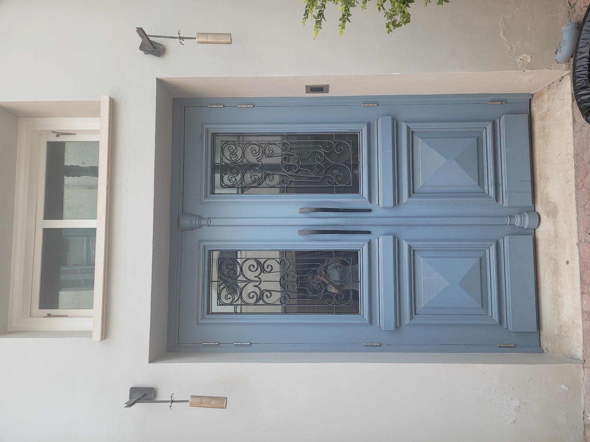 The image shows a light blue double door with decorative glass panels featuring wrought iron accents. On either side of the door, there are wall-mounted light fixtures with cylindrical, perforated shades. Above the door, a white-framed window with visible louvers behind the glass is present. The exterior wall is painted a light, neutral color. To the right of the door, a portion of a green plant is visible, and a grey PVC pipe curves towards the ground. The ground in front of the door is paved with concrete and brick. The scene appears to be an exterior entrance in Limassol, Cyprus.
