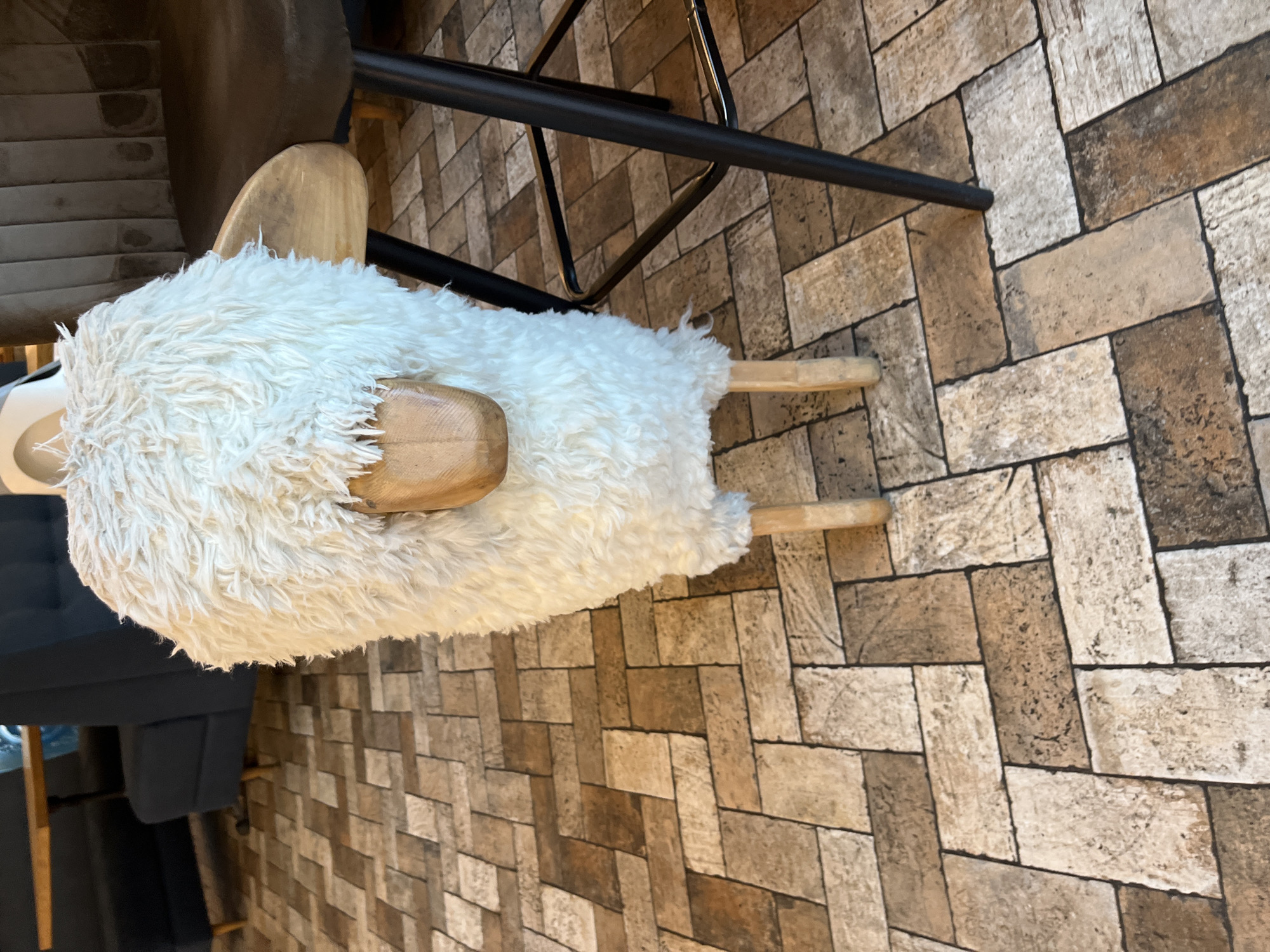 A fluffy white sheep-shaped ottoman sits on a patterned tile floor. The ottoman has wooden legs and a wooden face. To the right of the ottoman is a dark brown bar stool with a black metal base. In the background, dark blue banquette seating is visible, along with a wooden table. The environment appears to be an indoor public space such as a cafe or restaurant. The scene is not identifiable as being in Sochi, Russia.