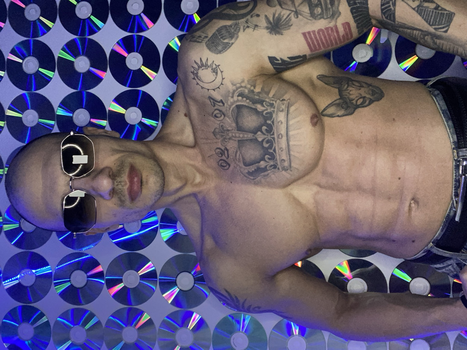 The image depicts a male individual, shirtless, facing the camera. His head is bald, and he wears dark, hexagonal-framed sunglasses. His lips appear slightly parted. He possesses a muscular physique with visible abdominal muscles.
Multiple tattoos are present on his upper body. On his chest, a large crown design is centered, with the numbers "02 07" tattooed above it. To the right of the numbers, on his right shoulder/pectoral area, are tattoos of a sun and a crescent moon. His right arm is covered with a full sleeve tattoo featuring various designs, including a microphone, palm trees, classic cars, a pyramid, and the word "WORLD" in red lettering. On his left side, below the pectoral muscle, a tattoo of a Sphynx cat head is visible. The lower portion of his torso shows he is wearing blue jeans secured by a black belt with a silver buckle.
The background consists of a wall entirely covered with numerous compact discs (CDs). These CDs reflect various hues, predominantly blue, purple, and green, suggesting the presence of artificial lighting. The specific location within Pleven, Bulgaria, is not identifiable from the visual information provided.