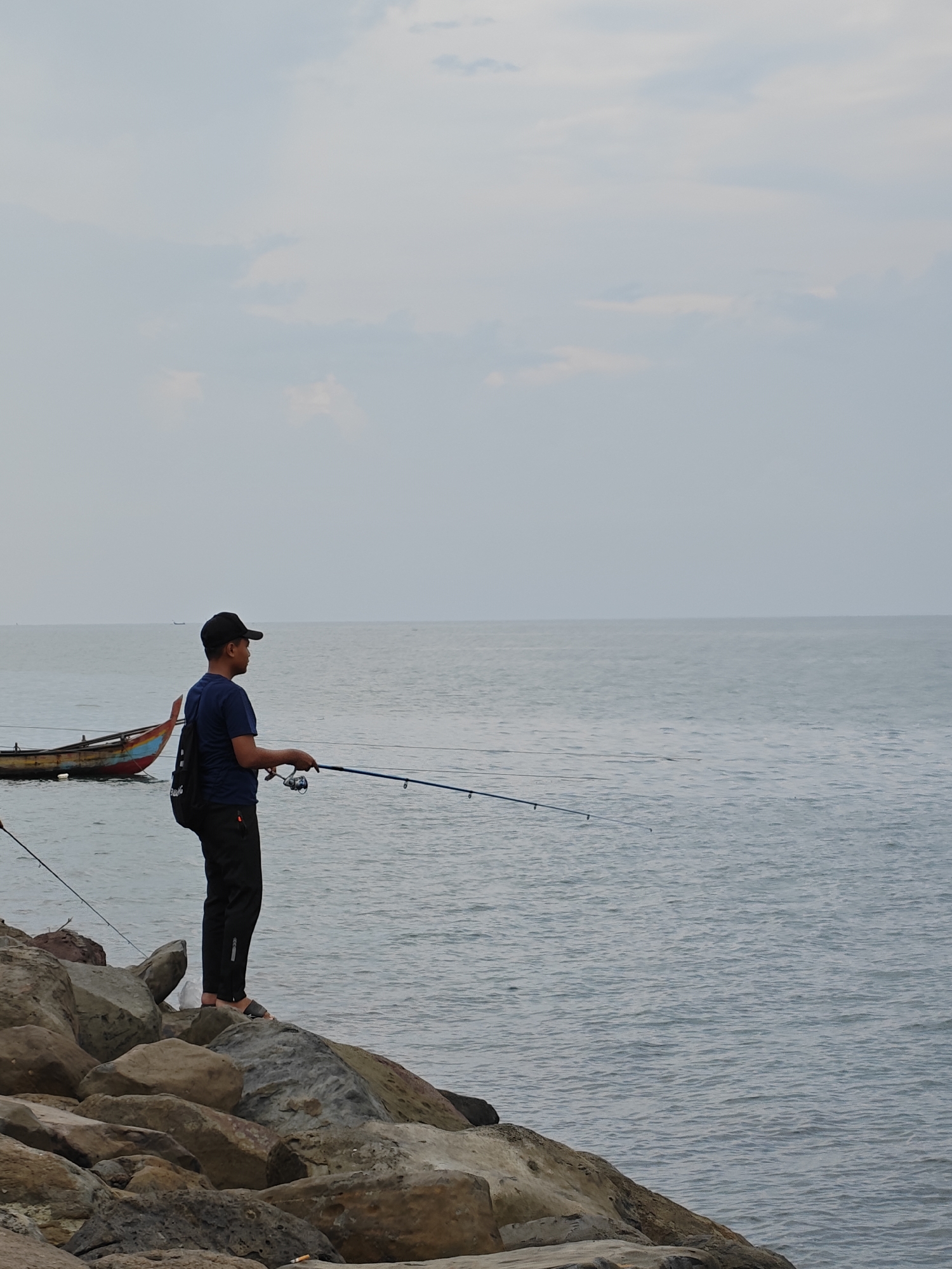 A man is captured fishing from a rocky breakwater along a calm sea under an overcast sky. He stands on a large, multi-toned rock, facing right and looking intently out over the water, holding a fishing rod with a reel. He is dressed in a dark blue t-shirt, black pants, a black baseball cap, and sandals, with a black backpack slung over his left shoulder that features partially visible text, possibly "SANCTUARY".
The foreground consists of a rugged array of large, weathered rocks forming the coastal edge. Beyond this, the tranquil grey-blue sea extends towards a soft horizon line, where it meets a uniformly light, cloudy sky. In the mid-ground, two small, traditional wooden boats are visible on the water; one is partially obscured on the far left, while the other, with vibrant red, blue, and yellow painted accents, is more distinct. Another fishing rod, possibly belonging to a different angler or a secondary setup, is partially visible in the bottom left corner.
The scene suggests an ordinary daytime activity in a coastal setting. The overcast weather creates soft, diffused lighting with no harsh shadows, contributing to a serene atmosphere. The man appears focused and calm while engaged in fishing. This peaceful coastal scene is set in Lhokseumawe, Indonesia, characterized by its rocky shoreline and traditional maritime elements.