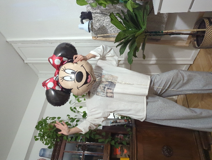 The image displays an indoor scene within an apartment room in Moscow, Russia. A single individual, positioned centrally, has their head obscured by a large, inflated Minnie Mouse foil balloon, which depicts Minnie's face, black ears, and a red-and-white polka dot bow. The person wears an oversized, light-colored t-shirt featuring a multi-panel photographic print of several individuals, along with loose-fitting grey sweatpants. Their left hand is raised and open, in a waving gesture, while their right hand appears to hold the balloon in front of their face.
To the left, a dark wooden antique-style cabinet with glass doors is visible, displaying various objects inside and adorned with trailing green plants. Several other green potted plants are present throughout the room, including a tall plant with large leaves, possibly a Ficus lyrata, housed in a woven basket on the right side of the frame. The floor is made of light-colored wood, and the walls are white, with two white doors discernible in the background. On the far right, a small light-blue object and a distinct black object rest on a textured white surface, possibly part of a cabinet or shelf. The room is brightly illuminated.