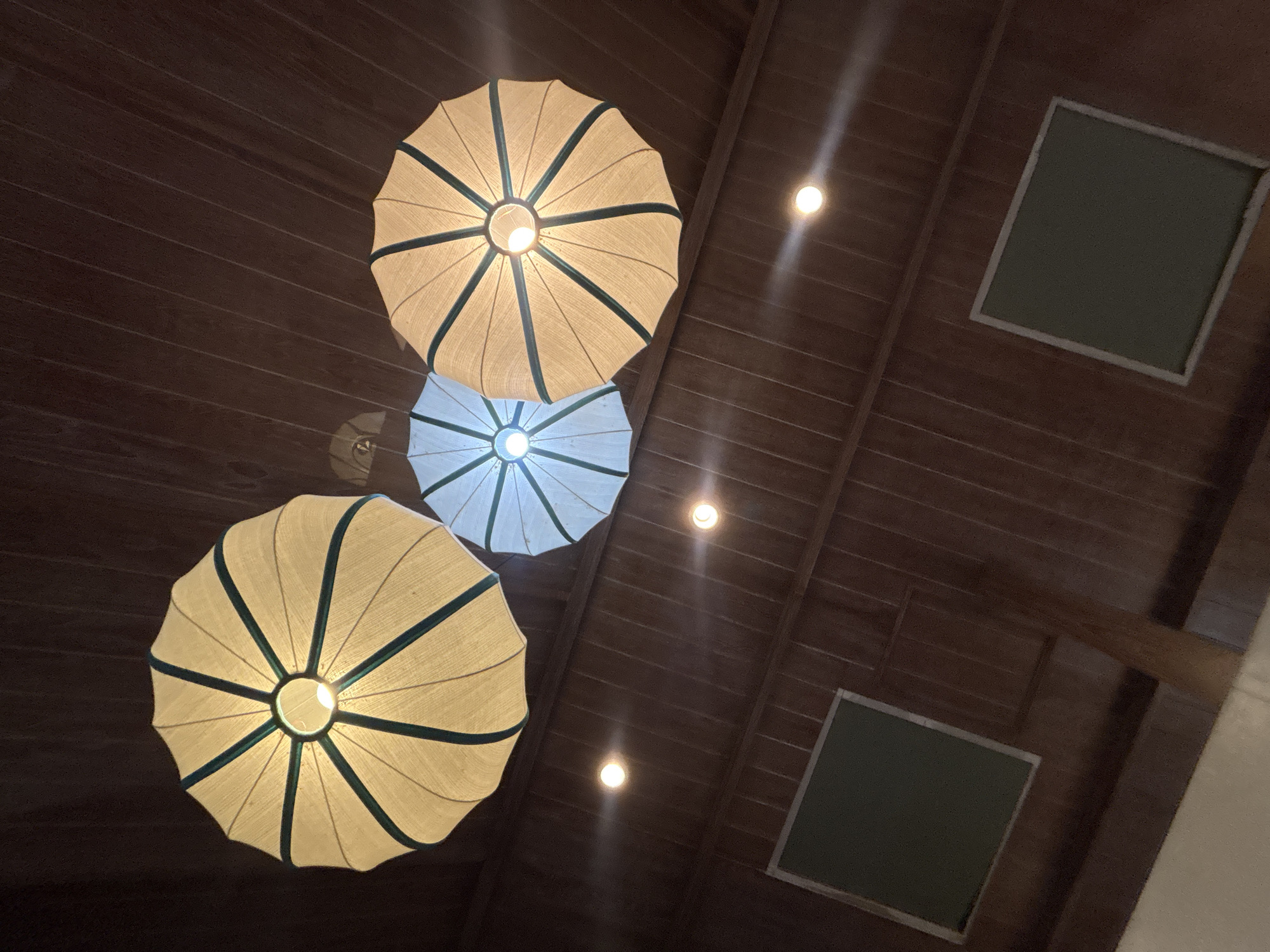 This is a low-angle shot of three suspended umbrella-shaped lanterns against a dark wooden paneled ceiling in Chalong, Thailand. The two outer lanterns are a creamy yellow with dark green trim, while the middle lantern is a pale blue with similar trim. All three are lit from within, casting a warm glow. Below the lanterns, two square, dark green framed panels are mounted on the wooden wall. Several small, bright circular lights are also visible on the ceiling, creating streaks of light. The scene appears to be indoors, likely a restaurant or covered seating area. There are no people or animals visible.