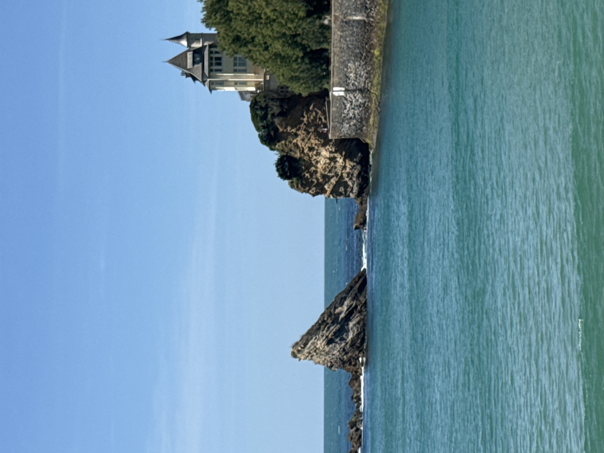 This image captures a scenic view of the Rocher de la Vierge (Virgin Rock) in Biarritz, France. In the foreground, the turquoise ocean displays gentle ripples under a clear blue sky. To the left, a prominent, jagged rock formation rises from the sea, with smaller rocky outcrops scattered around it. In the middle ground, a stone wall lines the edge of a cliff, separating the water from the land above. Perched atop the cliff is a distinctive building with multiple turrets and a light-colored facade, partially obscured by lush green trees. The building appears to be a historic structure, possibly a villa or a public building. The overall scene depicts a picturesque coastal landscape, bathed in natural sunlight.