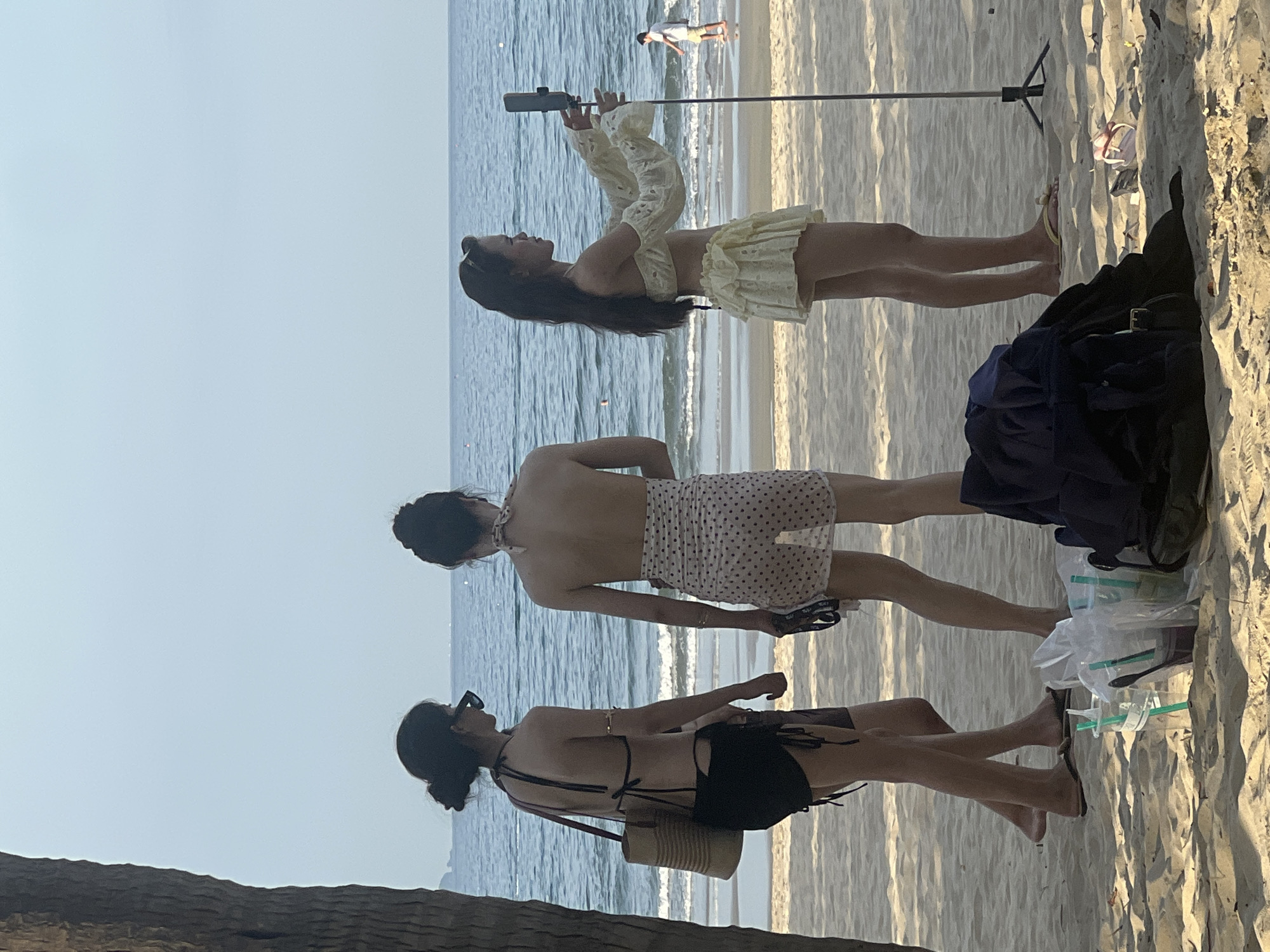 Three women are on a sandy beach in Đà Nẵng, Vietnam, with the ocean in the background. The woman on the left wears a black bikini and a straw bag. The woman in the middle wears a sheer polka-dot skirt over a bikini. The woman on the right wears a yellow off-the-shoulder top and a matching skirt, holding a smartphone mounted on a tripod. In the foreground, there are drinks in plastic cups and a pile of dark clothing. A man walks near the water in the distance.