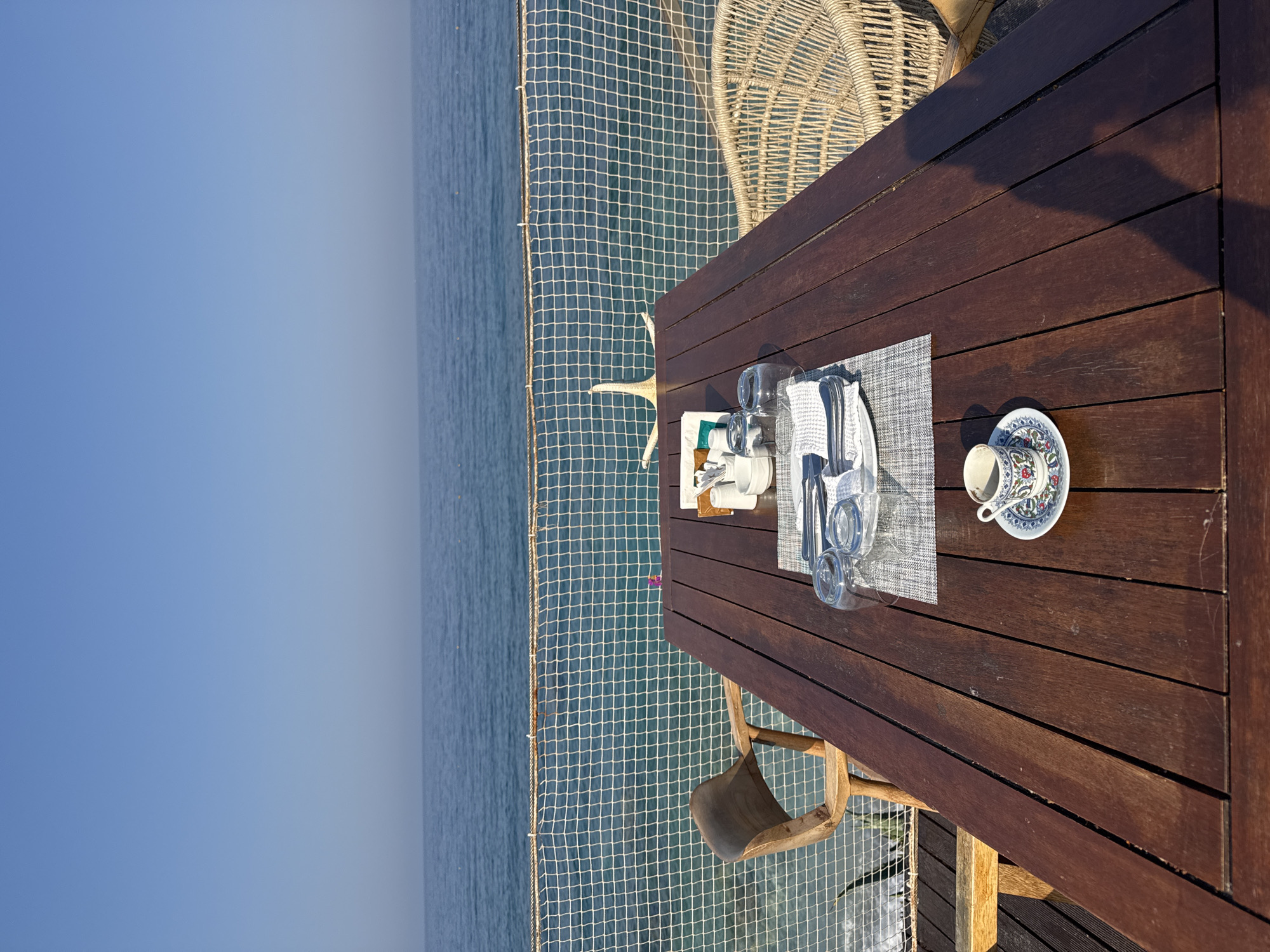 An outdoor dining setup for two is positioned on a wooden deck overlooking the sea in Fethiye, Turkey. The table is set with plates, cutlery, glasses, napkins, and a small coffee cup and saucer. Two chairs, one wooden and one rattan, are placed at the table. A netting barrier separates the deck from the water, and a starfish decoration is visible on the netting. The sea and sky are clear, suggesting a bright, sunny day.