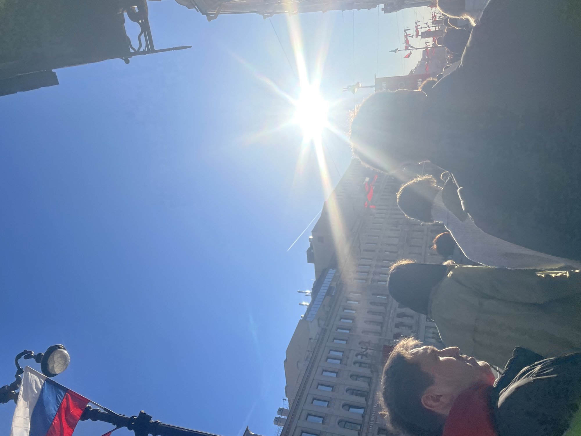 The image shows a crowd of people in Saint Petersburg, Russia, looking up at the sky. A Russian flag is visible on a streetlight in the foreground. The sun is shining brightly, and there is a tall building in the background. The crowd is likely gathered for an event or celebration. The building appears to be an older structure with many windows. The sky is clear and blue.
