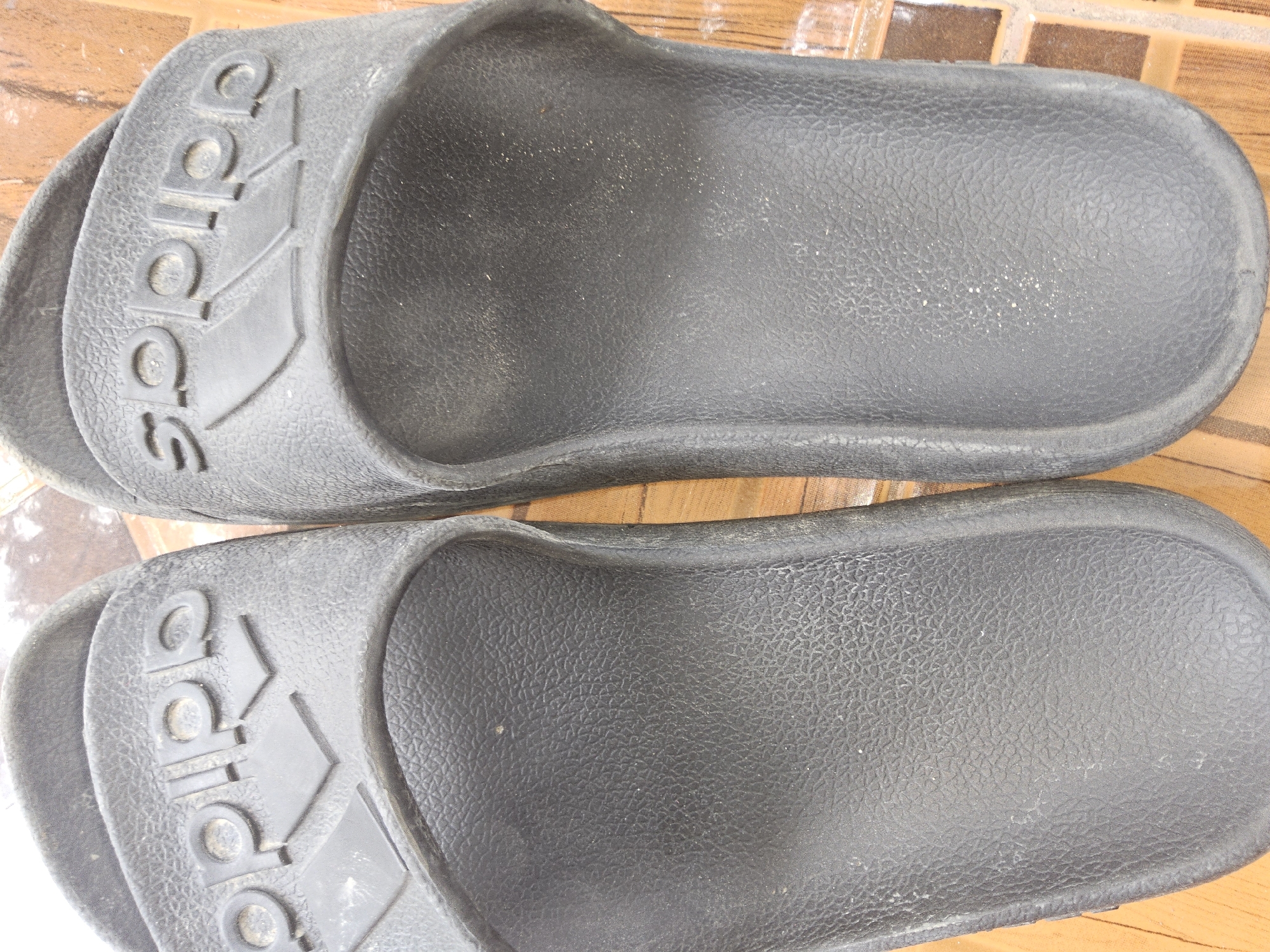 This is a close-up, top-down shot of two black Adidas slides on a wooden surface. The slides are positioned side-by-side, with their soles facing upwards. The Adidas logo, consisting of the word "adidas" and three parallel stripes, is clearly visible on the top strap of each slide. The surface of the slides appears textured and slightly dusty. The wooden surface beneath the slides has a tile-like pattern with visible grout lines. The lighting is diffuse, casting no harsh shadows. The image is focused on the slides and does not contain any elements that indicate a specific location within Payagaboh, Indonesia, or any context beyond the objects themselves.