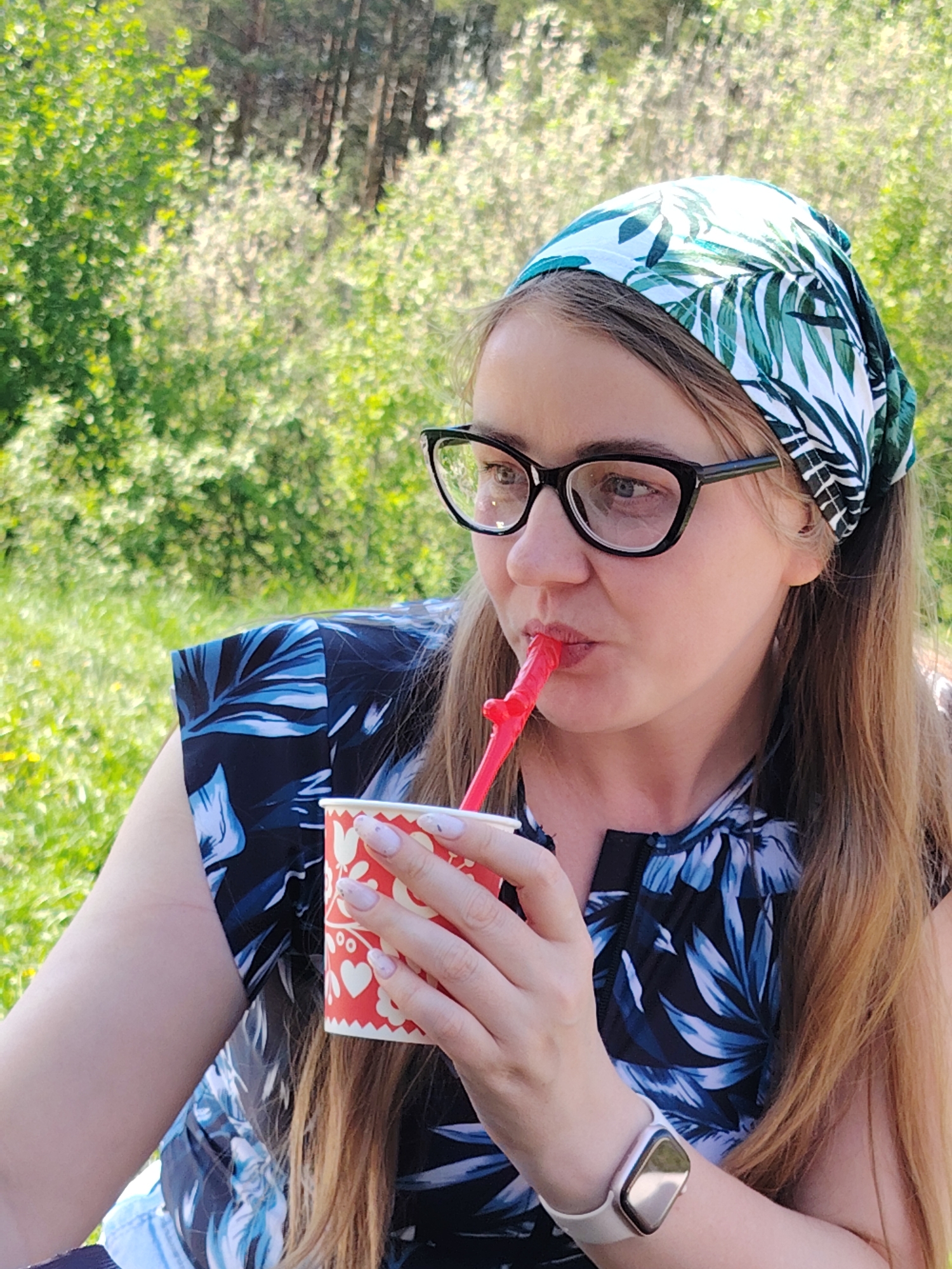 A woman sits outdoors in a park or wooded area, possibly in Chaikovsky, Russia. She wears a headscarf patterned with green and white tropical leaves, black-framed glasses, and a dark blue top with a floral pattern. She is drinking from a red paper cup with white hearts, using a red drinking straw shaped like a novelty object. She wears a light-colored watch on her left wrist. The background includes green trees and foliage, with dappled sunlight filtering through the leaves.